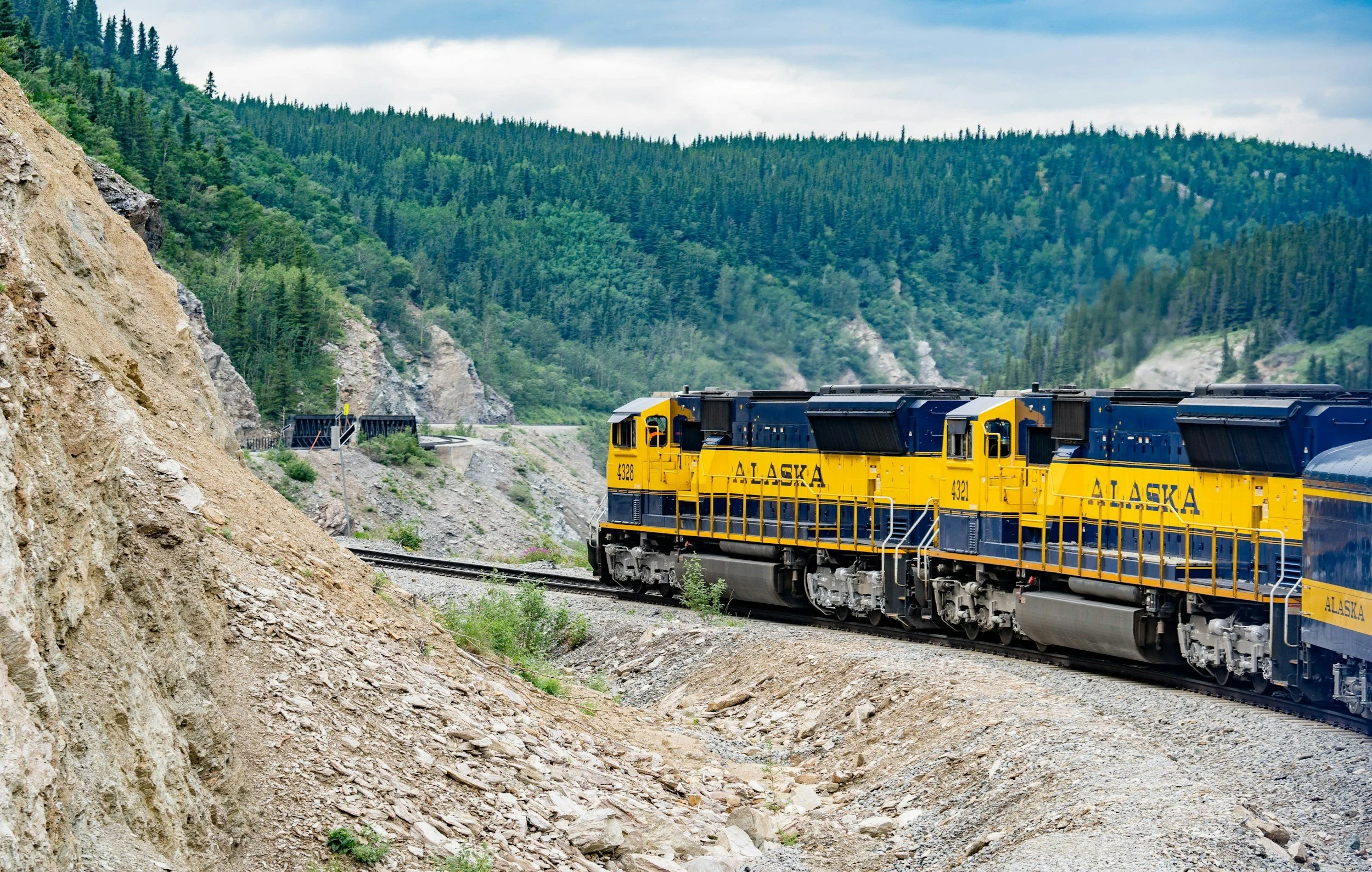 A yellow and blue Alaska train traveling through a mountainous landscape with green pine trees and rocky terrain under a cloudy sky.