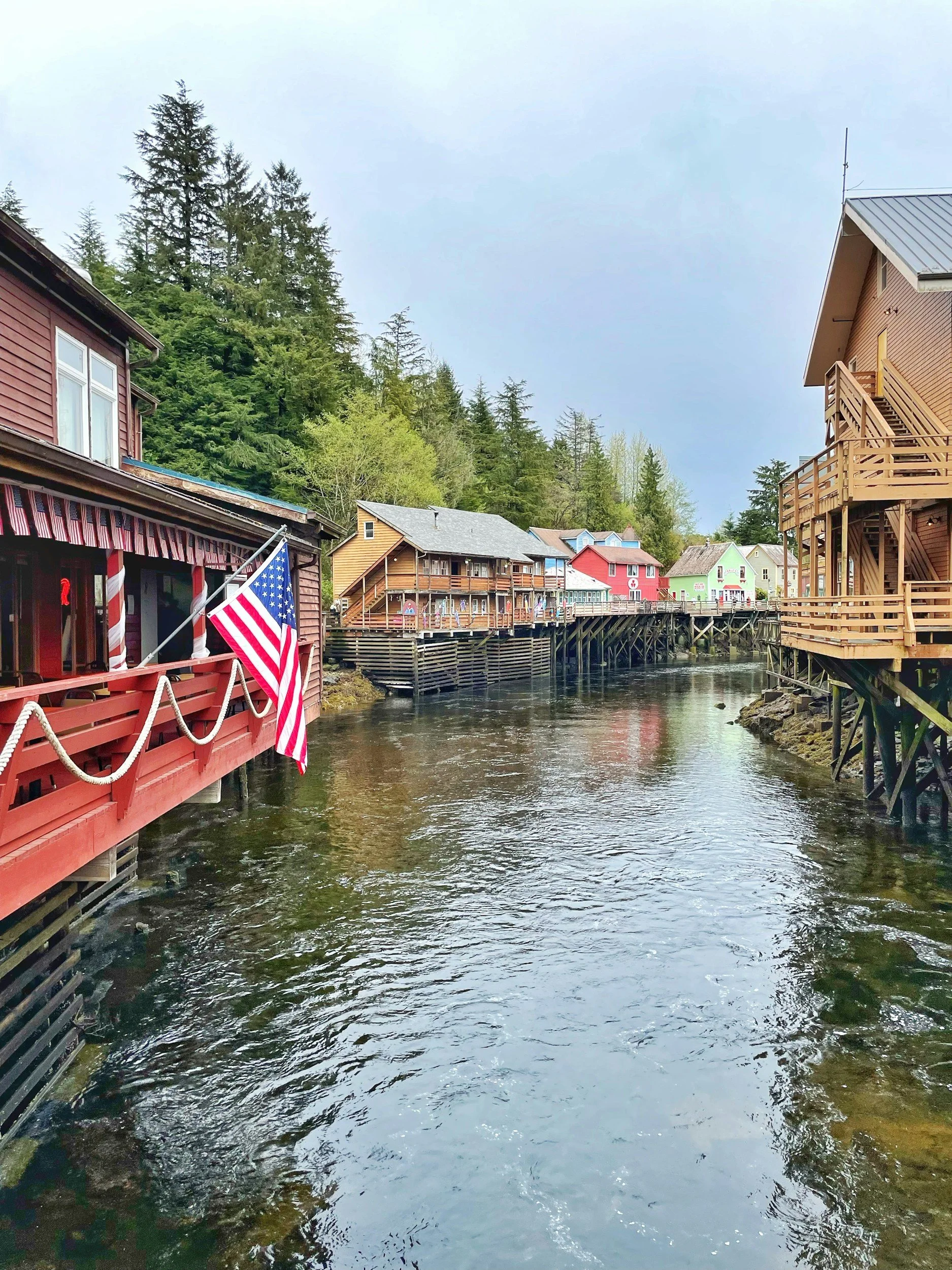 Colorful houses on stilts along a river, with trees and a cloudy sky in the background.