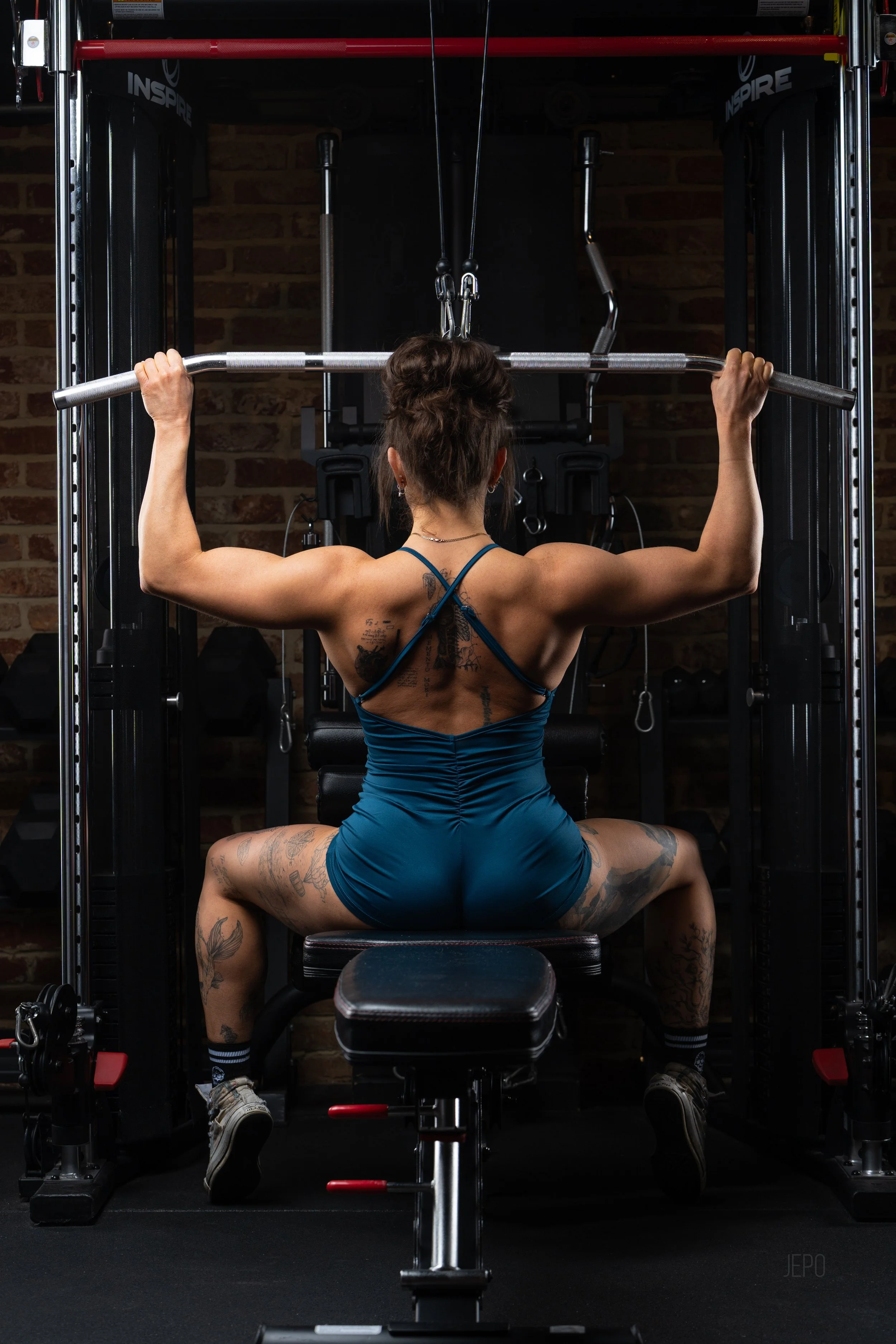 A woman with tattoos performing a lat pulldown exercise in a gym, seated with her back toward the camera, gripping a wide bar overhead.