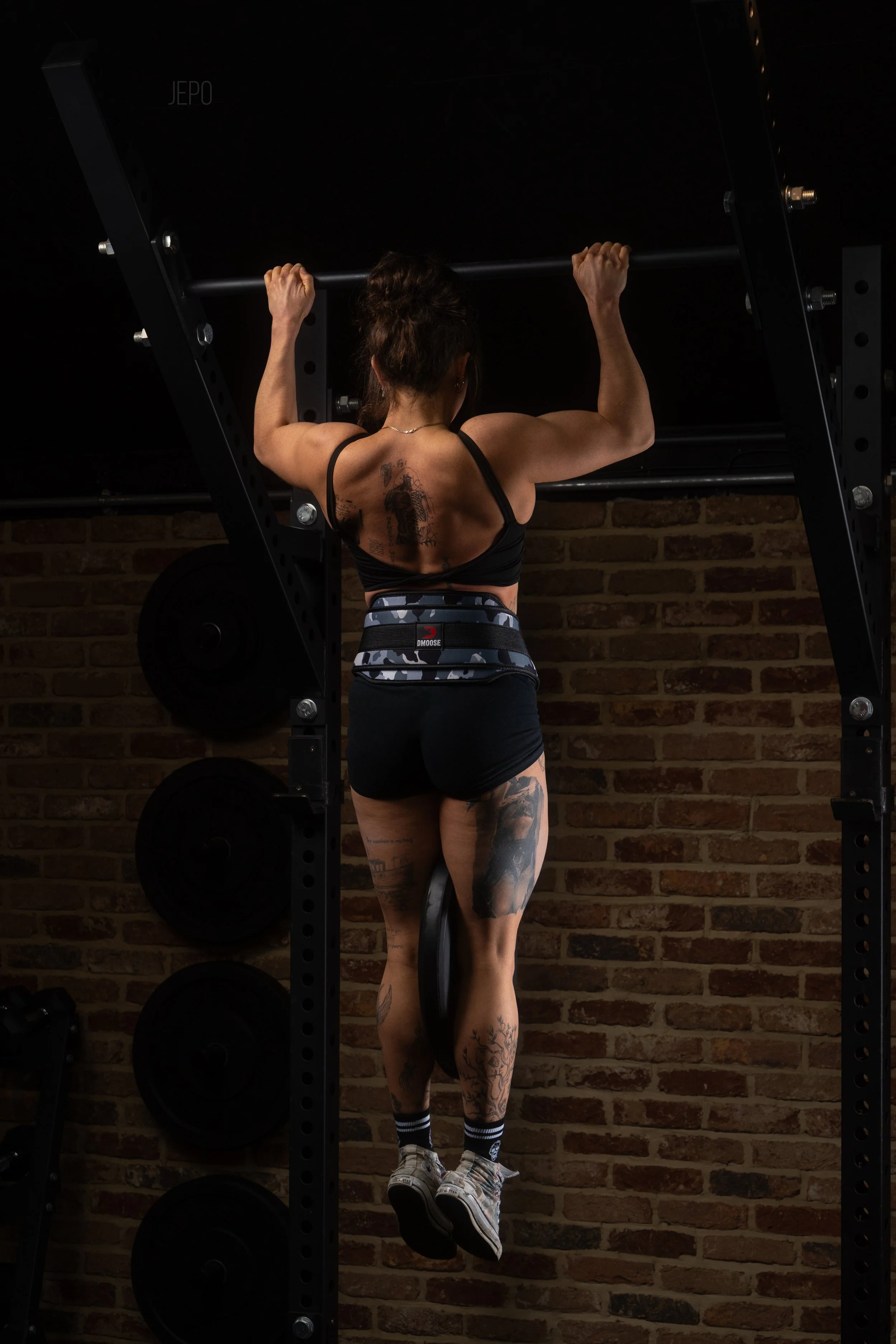 A woman with tattoos is doing a dip exercise on a pull-up bar in a gym with a brick wall background.