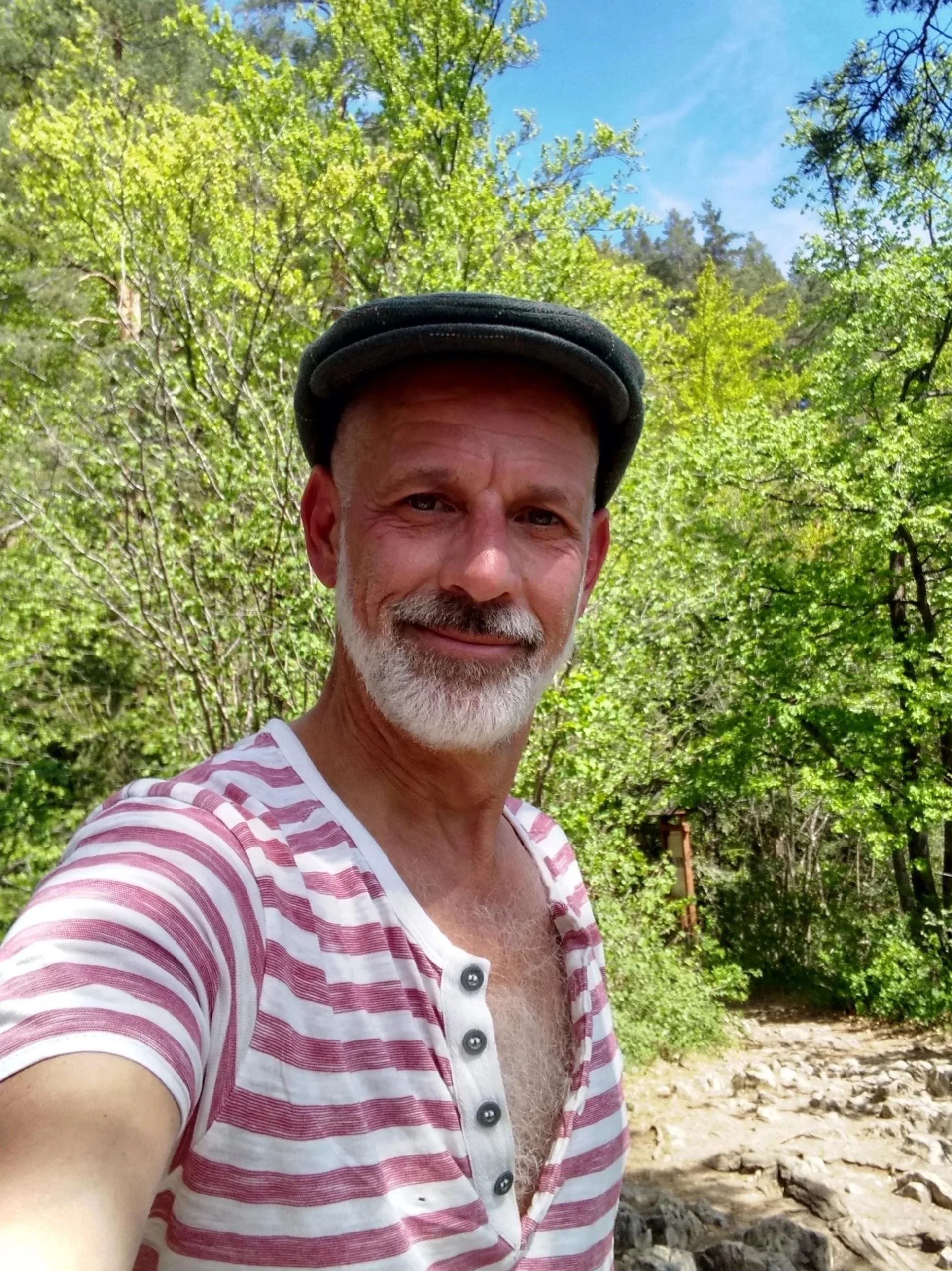 A middle-aged man with a gray beard and mustache, wearing a black cap and a red and white striped shirt, taking a selfie outdoors in a lush green forest with a small creek or river in the background under a partly cloudy sky.