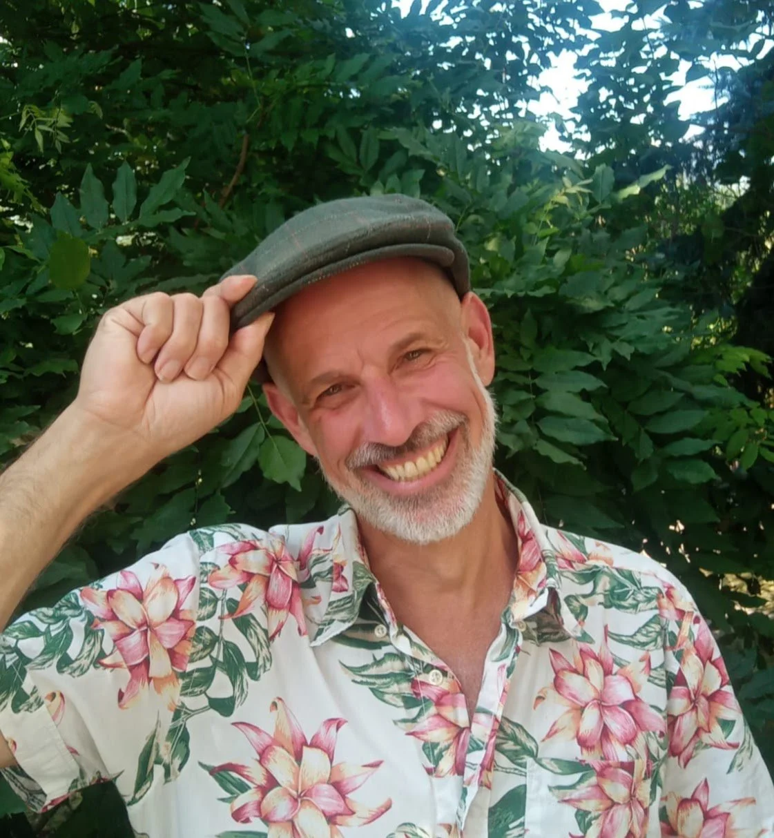 A smiling man with a beard wearing a floral shirt and a hat, posing outdoors in front of green foliage.