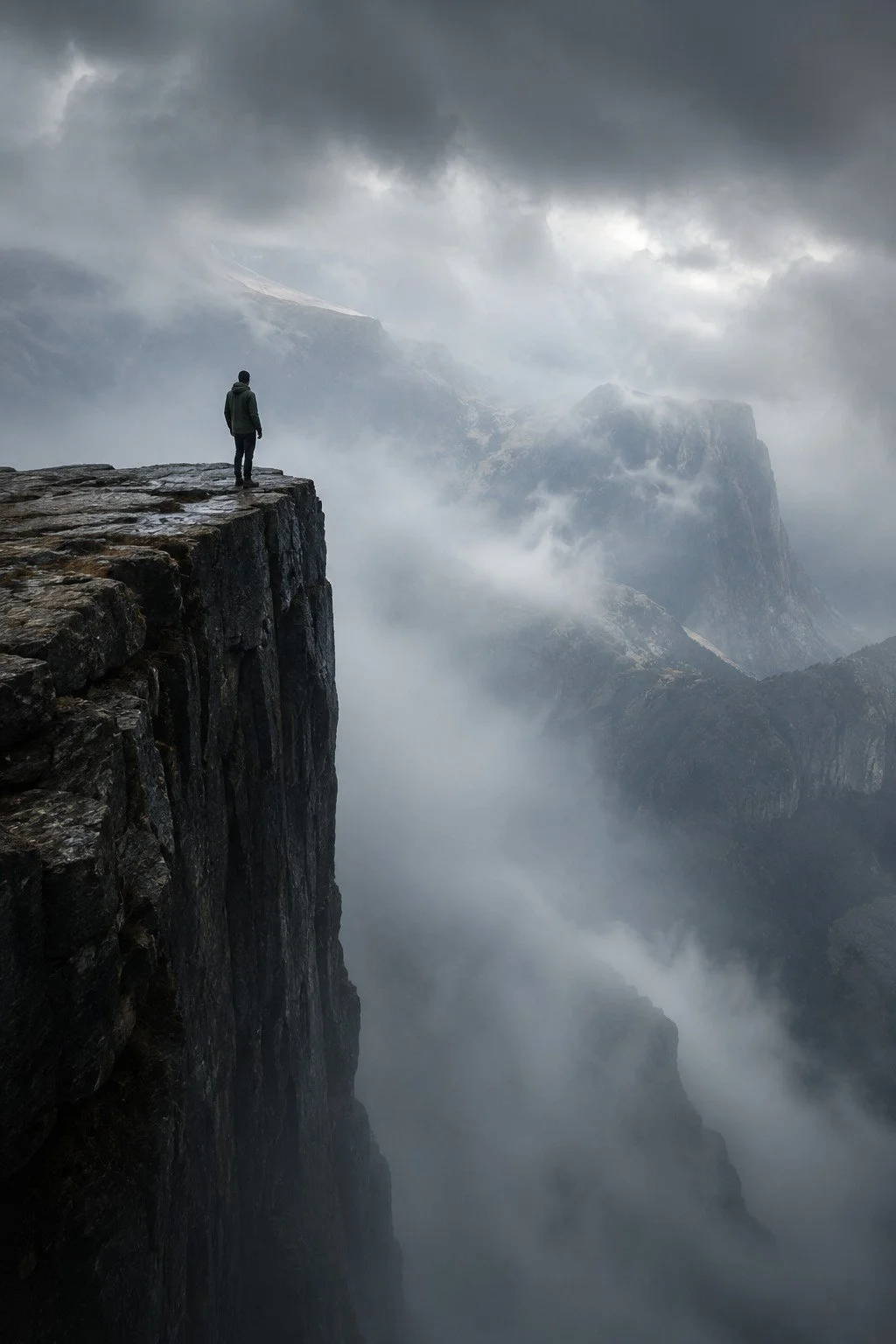 A person standing on the edge of a rocky cliff overlooking a foggy, mountainous landscape with clouds and mist surrounding the peaks.