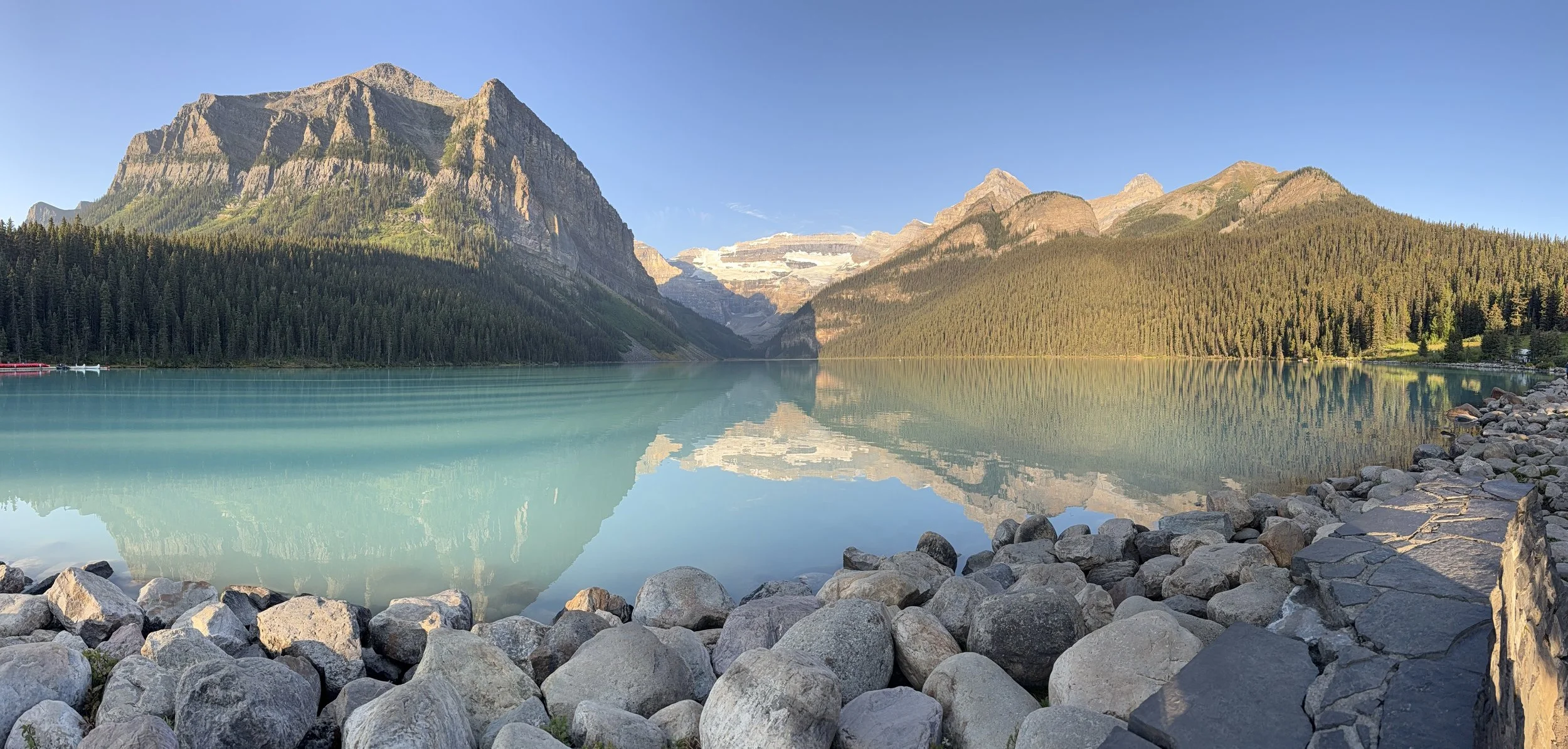 Scenic view of a calm turquoise lake surrounded by rocky shoreline and dense green forests, with rugged mountains in the background under a clear blue sky.