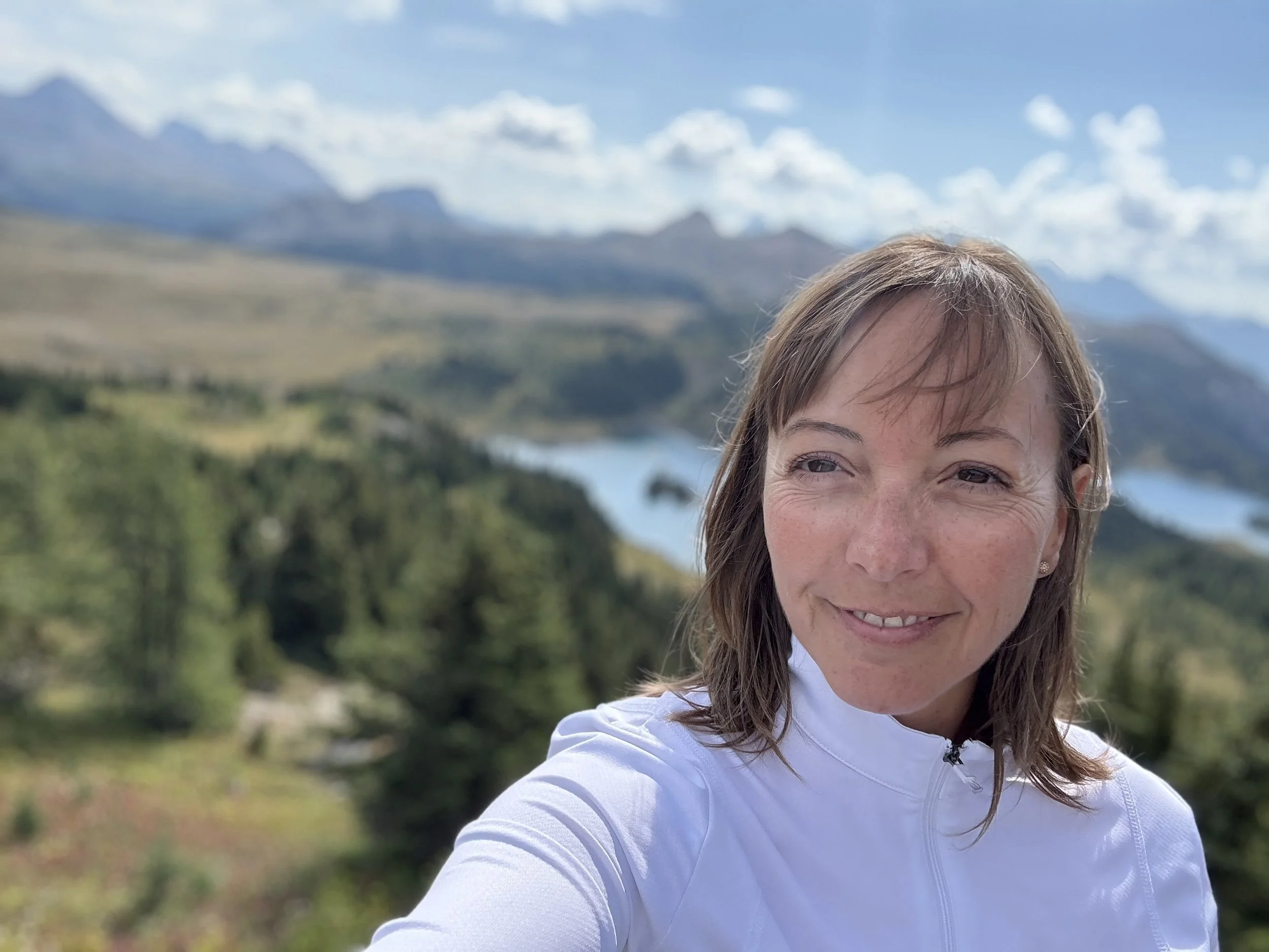 A woman with light brown hair and freckles smiling outdoors in front of mountains and a cloudy sky.