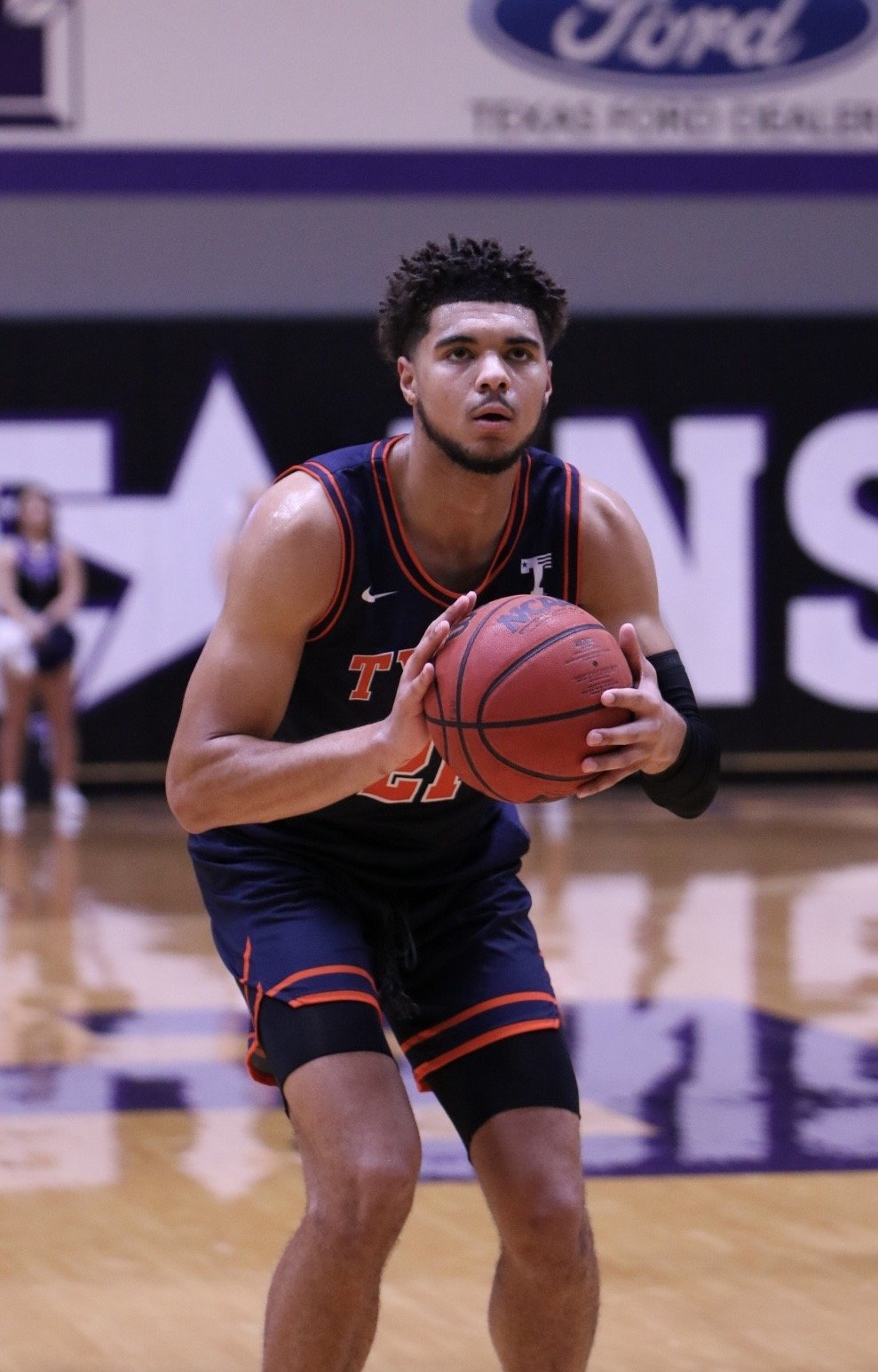 A young male basketball player wearing a black and orange uniform, making a free throw shot on an indoor basketball court.