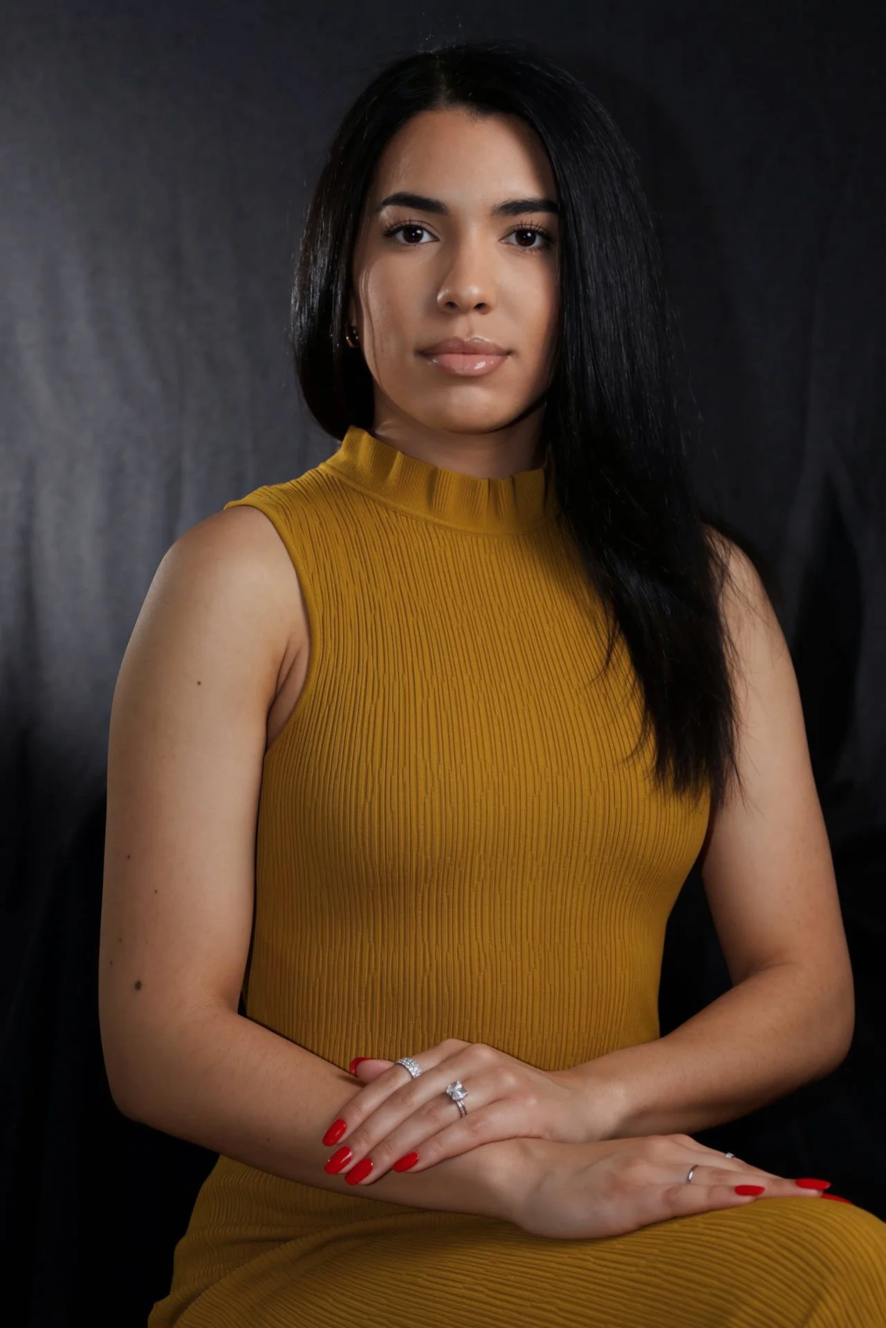 Portrait of a woman with long dark hair wearing a sleeveless mustard yellow top, with jewelry including rings and earrings, against a dark background.