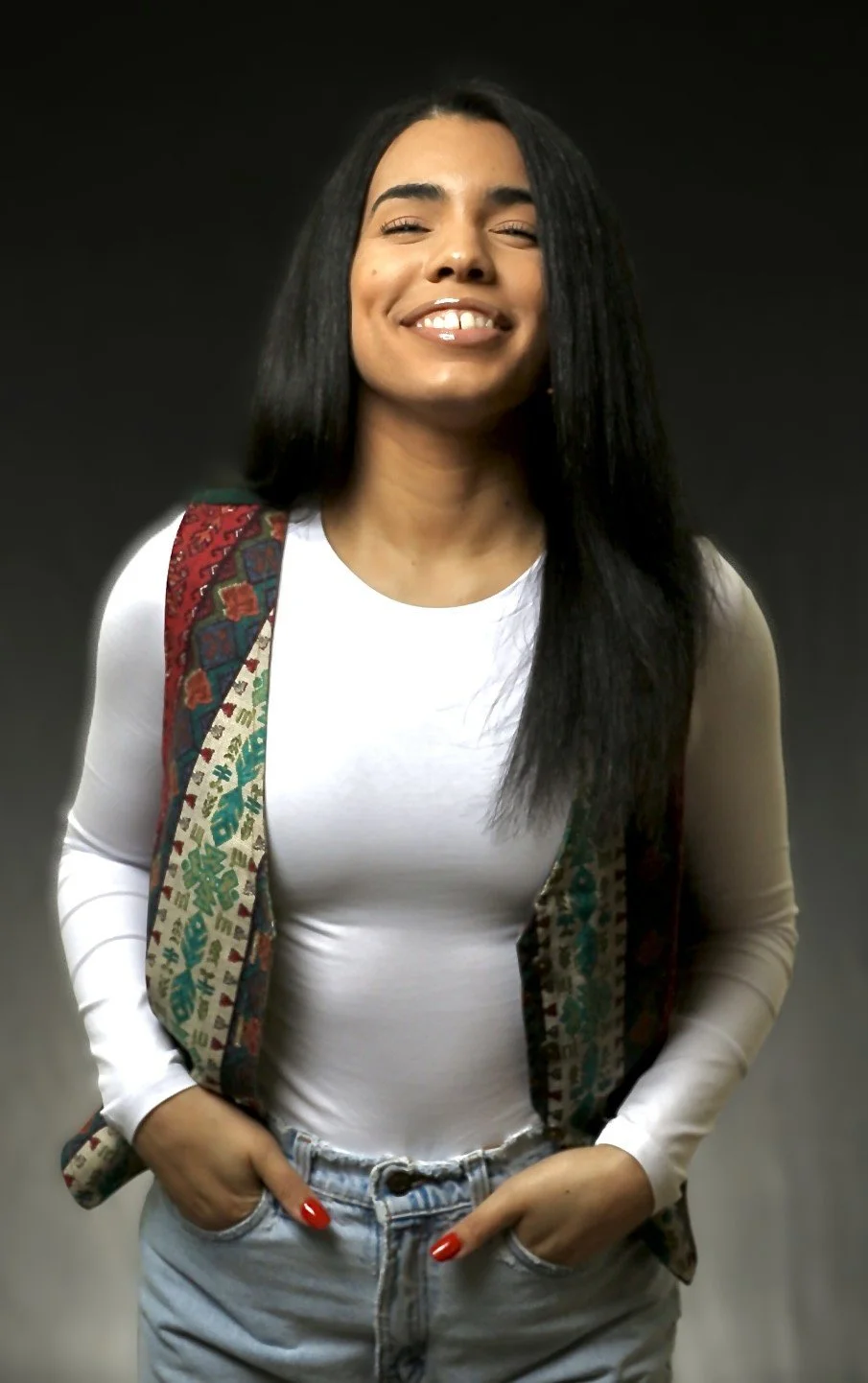 A young woman with long black hair smiling, wearing a white long-sleeve top, a patterned vest, and jeans, standing against a dark background.