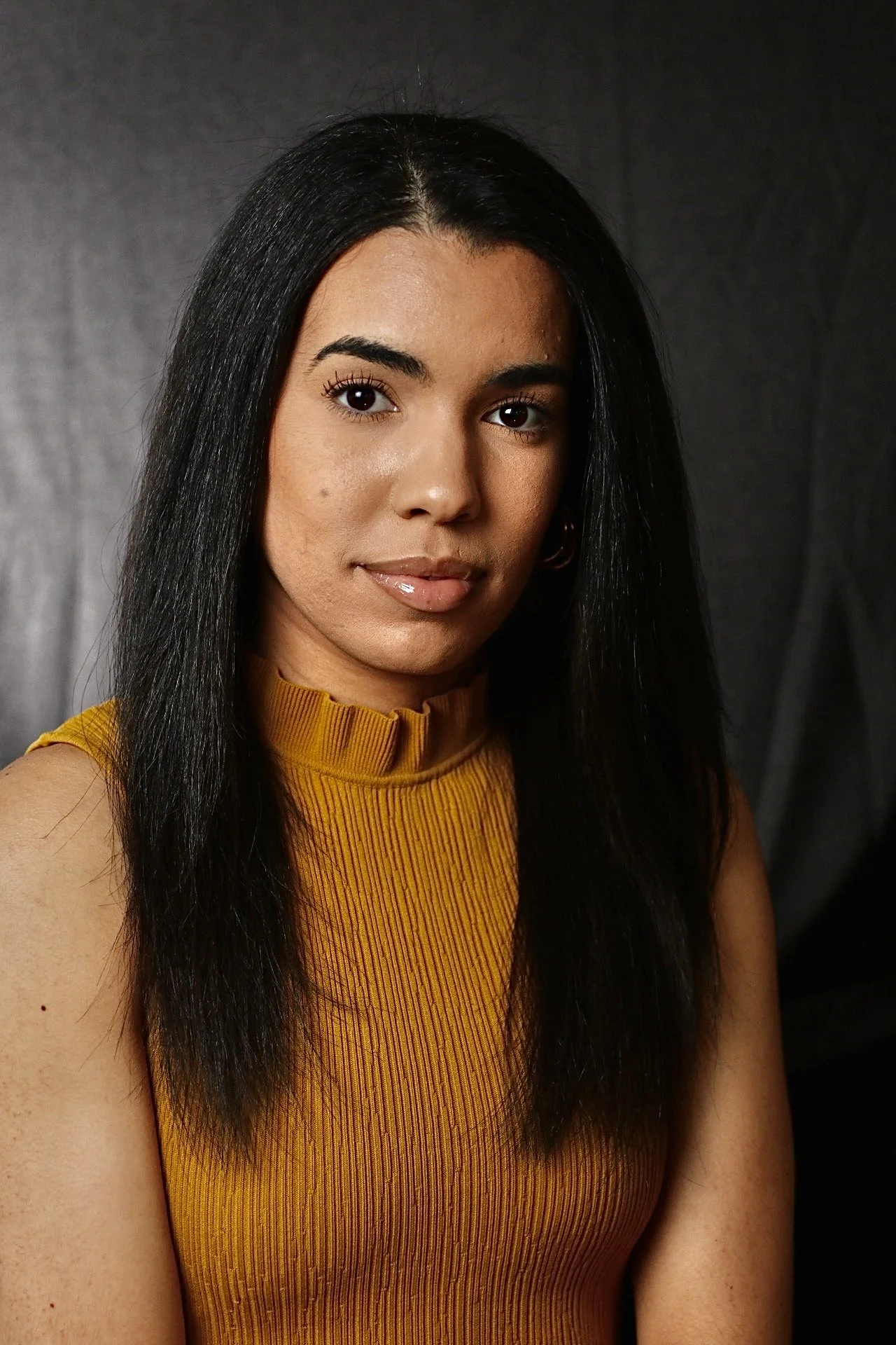 Close-up portrait of a woman with long black hair, wearing a mustard yellow sleeveless top, against a dark background.