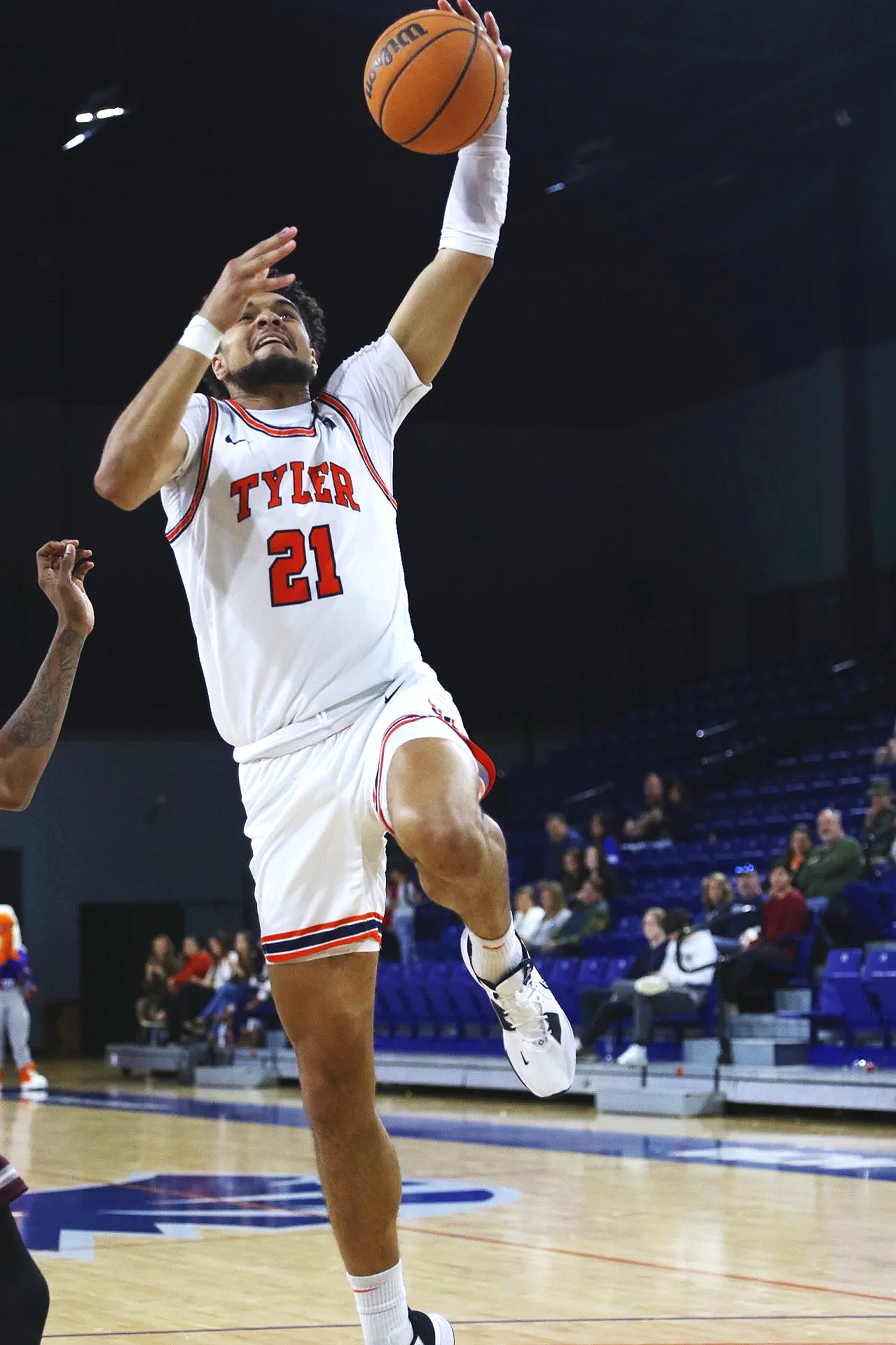 A basketball player wearing a white jersey with red and black trim, number 21, is jumping in the air to make a shot, holding an orange basketball with one hand. The player is wearing white socks and white sneakers with black accents, and a white arm sleeve. The background shows an indoor basketball court with spectators seated in blue chairs.