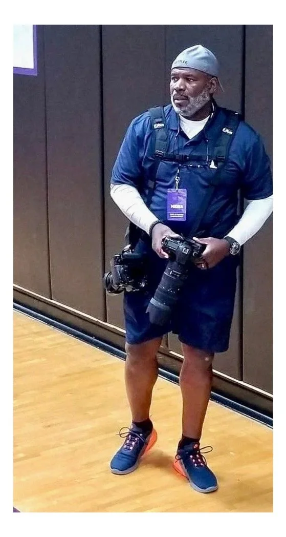 Man in athletic attire, wearing a gray hat, navy shirt, navy shorts, and running shoes, holding a professional camera with a large lens, standing indoors against a dark brown wall.
