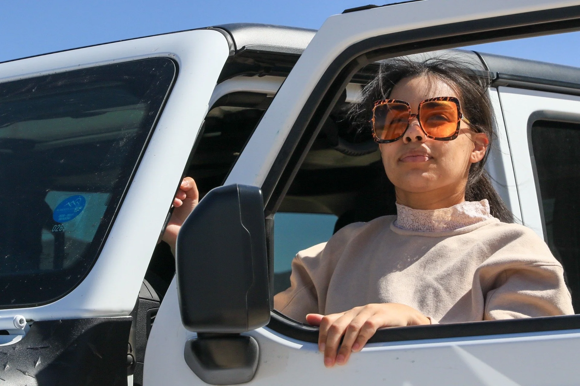 A woman with dark hair, wearing large tortoiseshell sunglasses and a beige high-collared blouse, sits in the driver's seat of a white vehicle, looking out of the open window with a slight smile. The sky is clear and blue in the background.