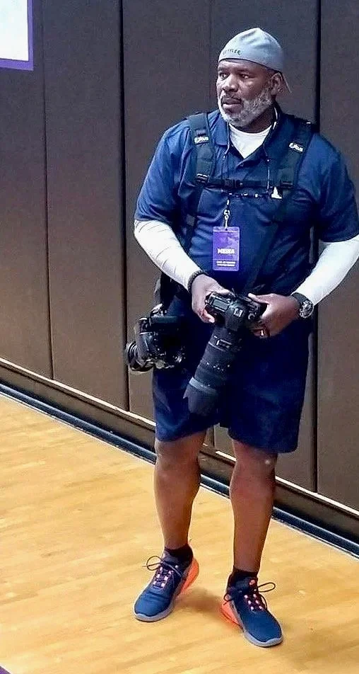 A man dressed in casual athletic wear, including a gray cap, navy shirt, white long sleeve underneath, navy shorts, and running shoes, holding a professional camera with a large lens, standing indoors against a dark wall.