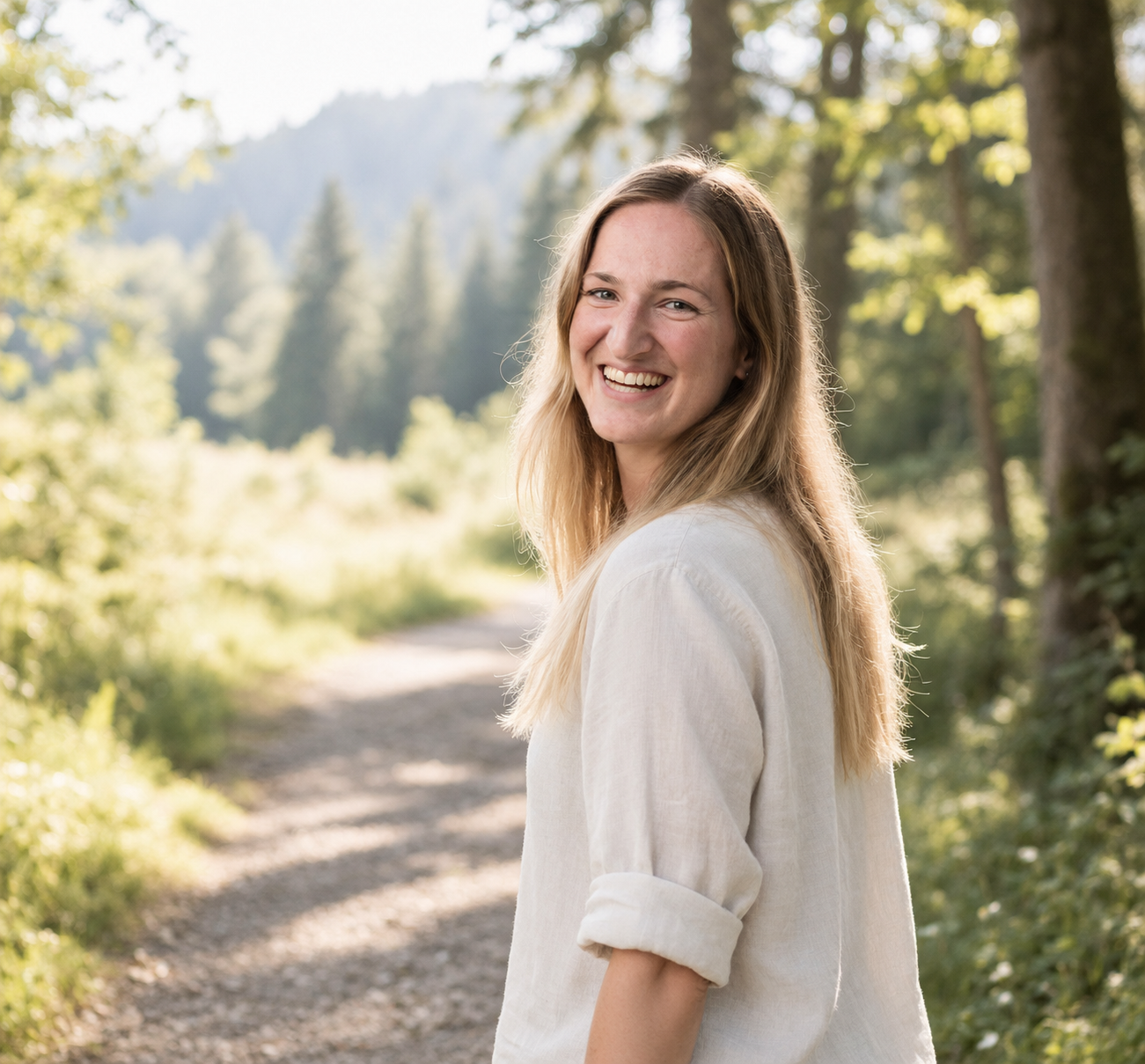 Junge Frau mit blonden Haaren, lächelnd, im weißen Hemd, auf einem Waldweg mit grünen Bäumen und Bergen im Hintergrund bei Sonnenlicht.