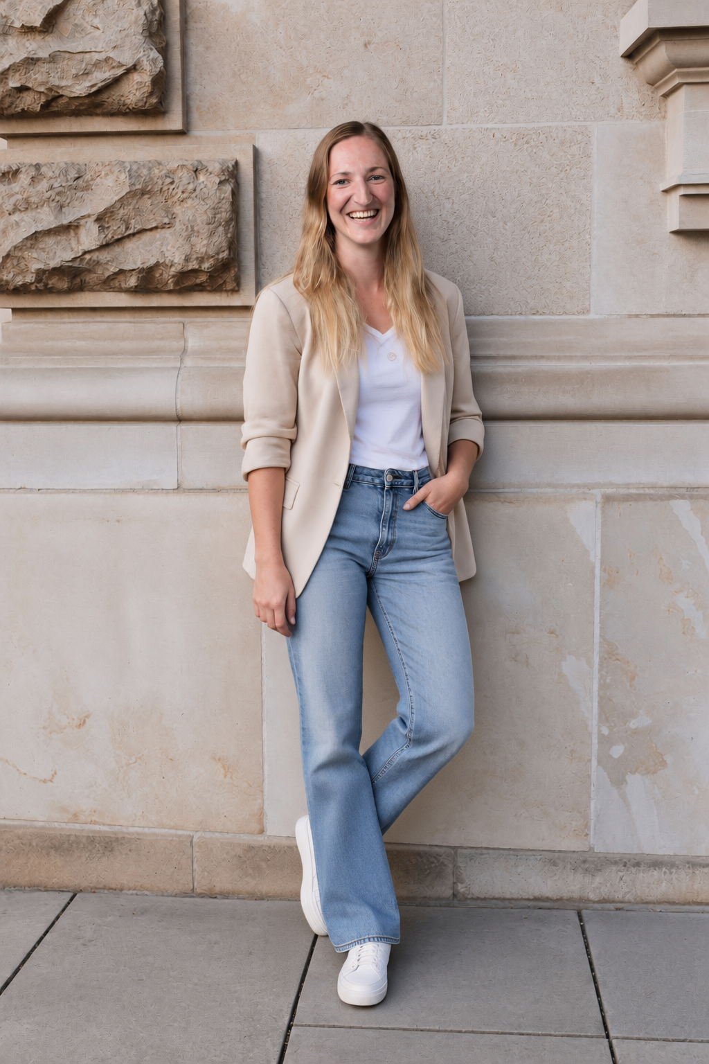 Freudestrahlende junge Frau in Jeans und Blazer lehnt an einer Steinmauer.