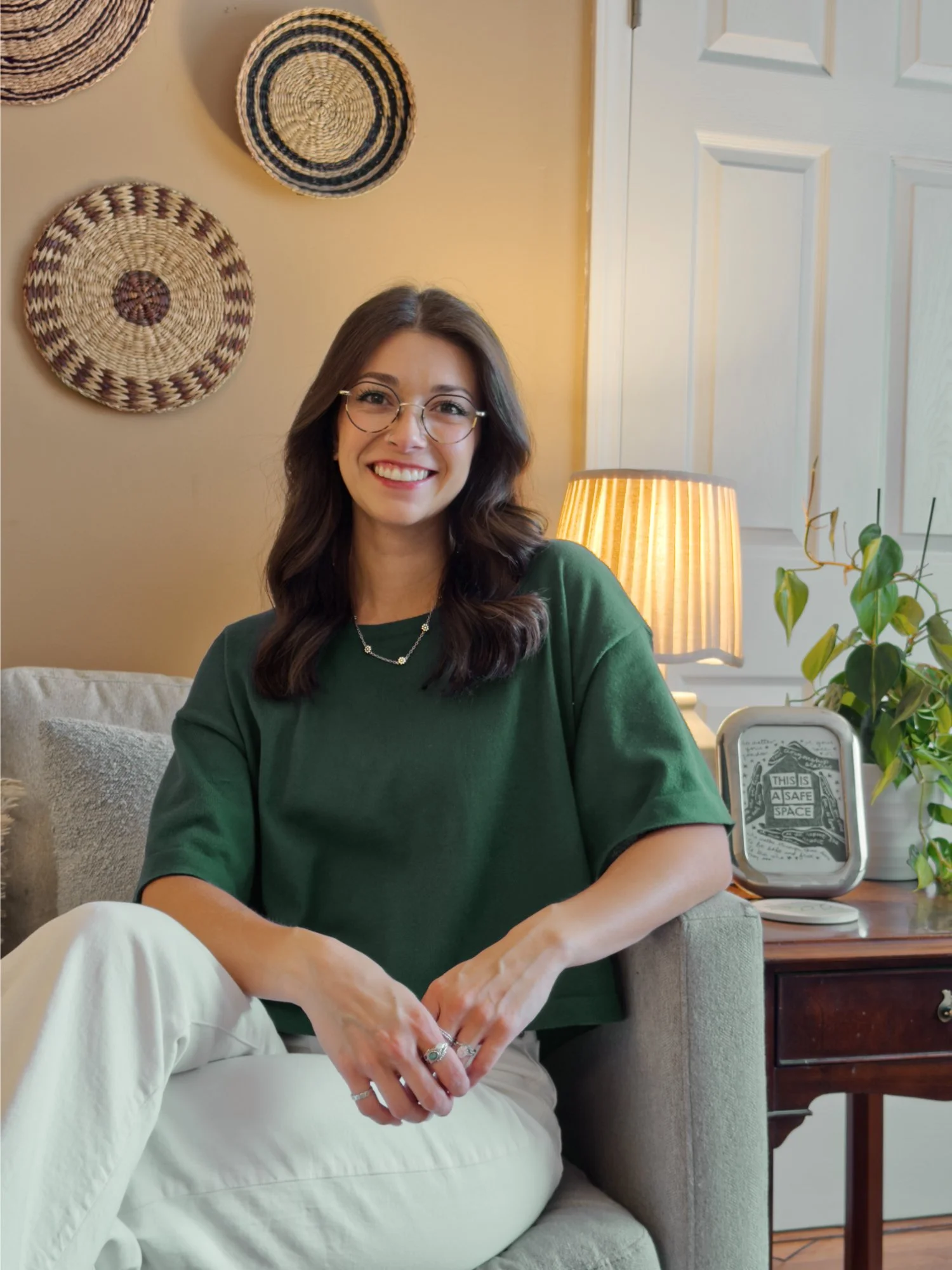 Gabby Lux, LPC, seated on the couch in her Greenville therapy office, smiling beside a lamp and plants.