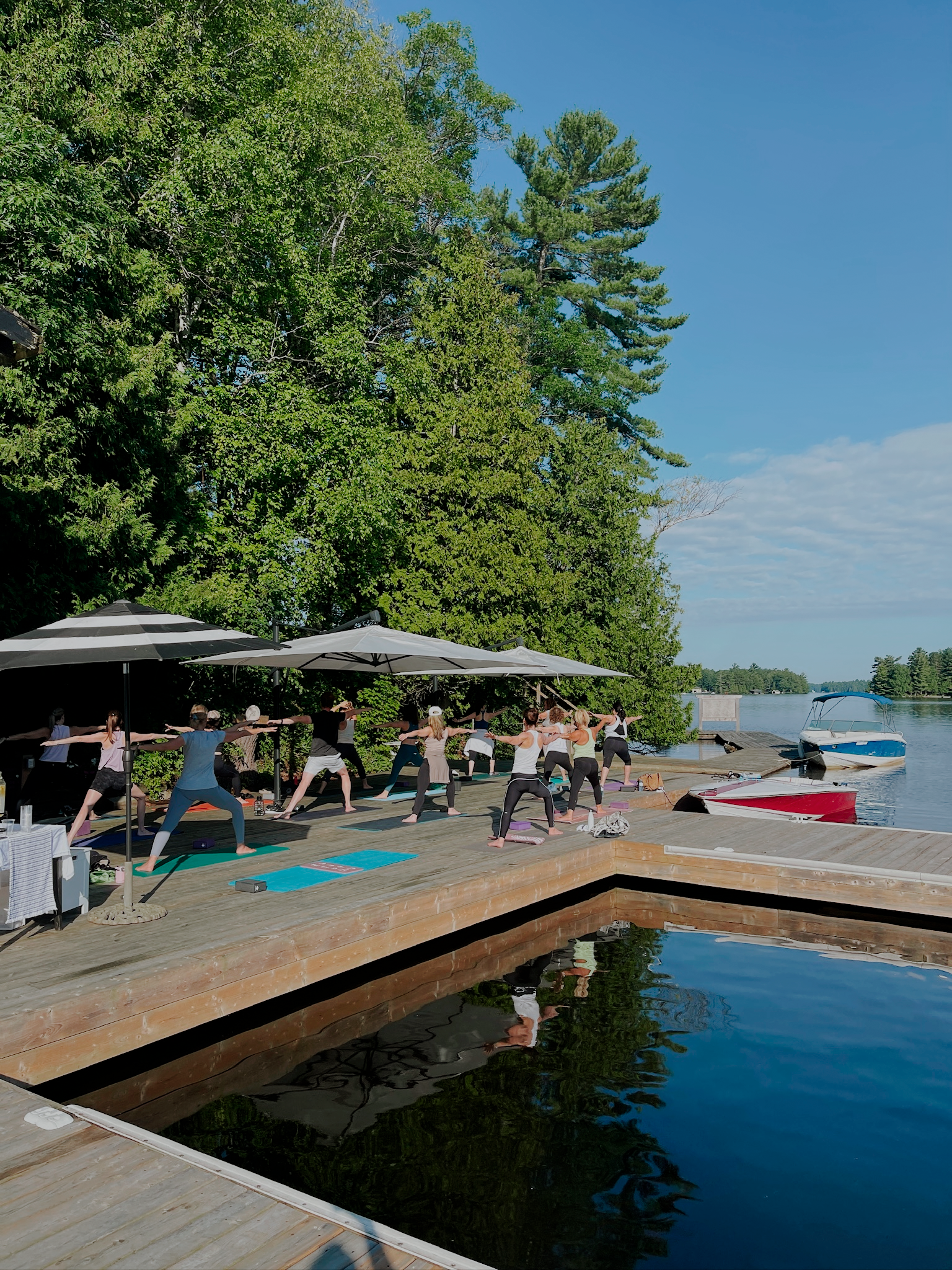 A group of people practicing yoga on a wooden dock by a lake, surrounded by trees and under umbrellas on a sunny day.