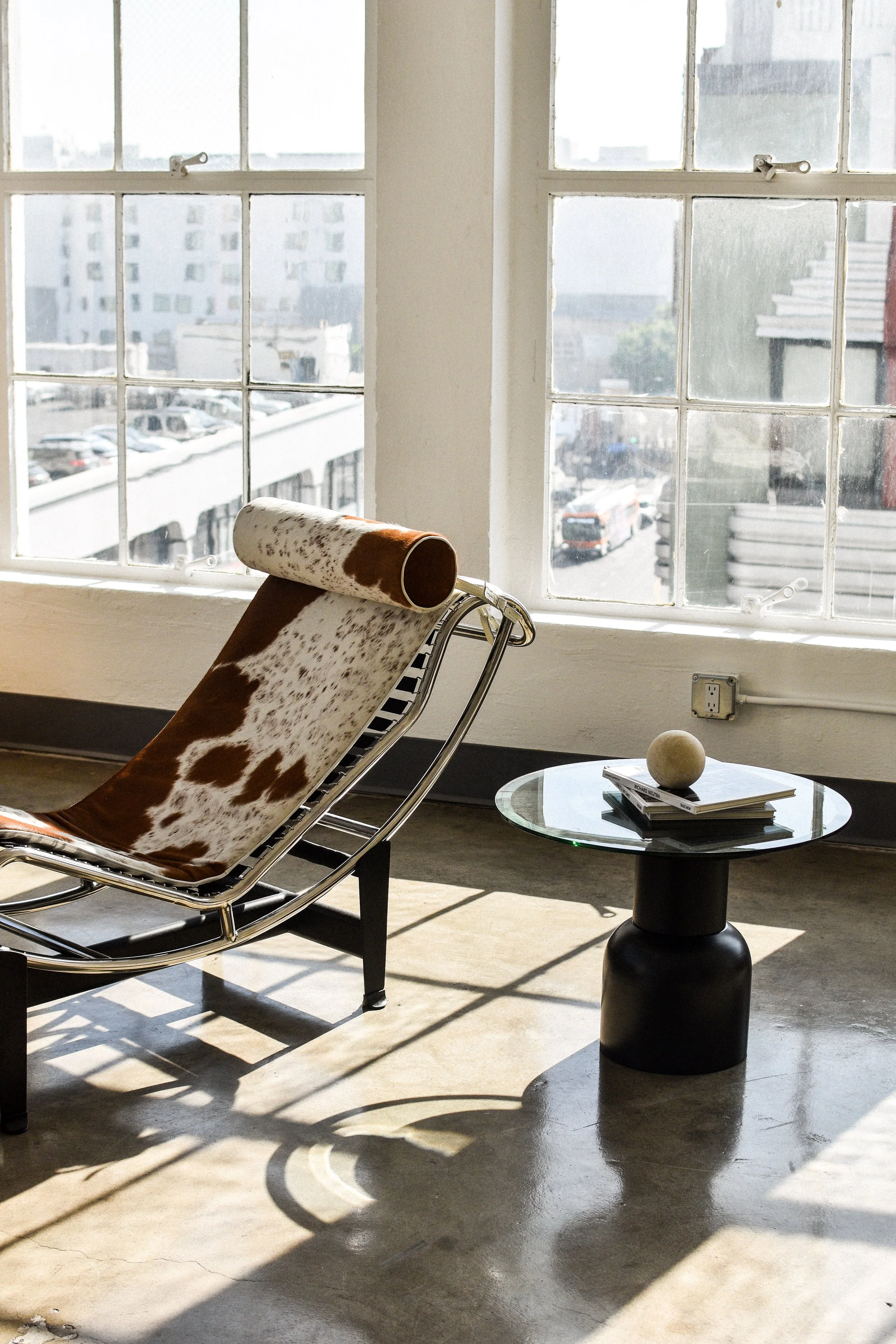 A cowhide chaise lounge chair with a chrome frame next to a black side table with a white sphere, placed on a concrete floor by large window panes with a city view in the background.