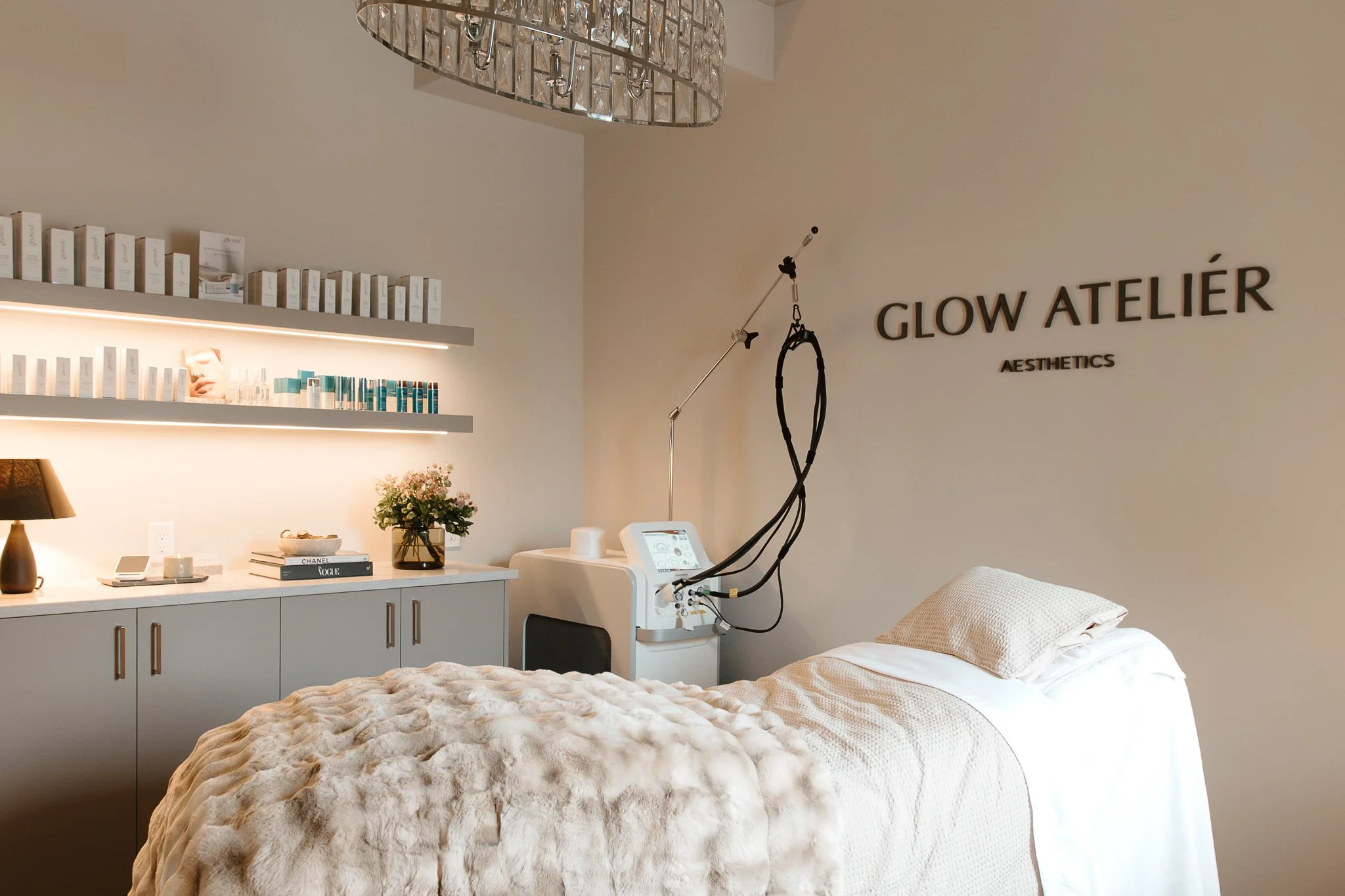 A cozy examination room with a bed covered in a plush beige blanket and a pillow, a white cabinet with decorative books and a flower arrangement, a bedside lamp, medical equipment including a device with attached black cords, and shelves with skincare products. The wall features the sign 'GLOW ATELIER AESTHETICS' and a stylish chandelier hangs from the ceiling.