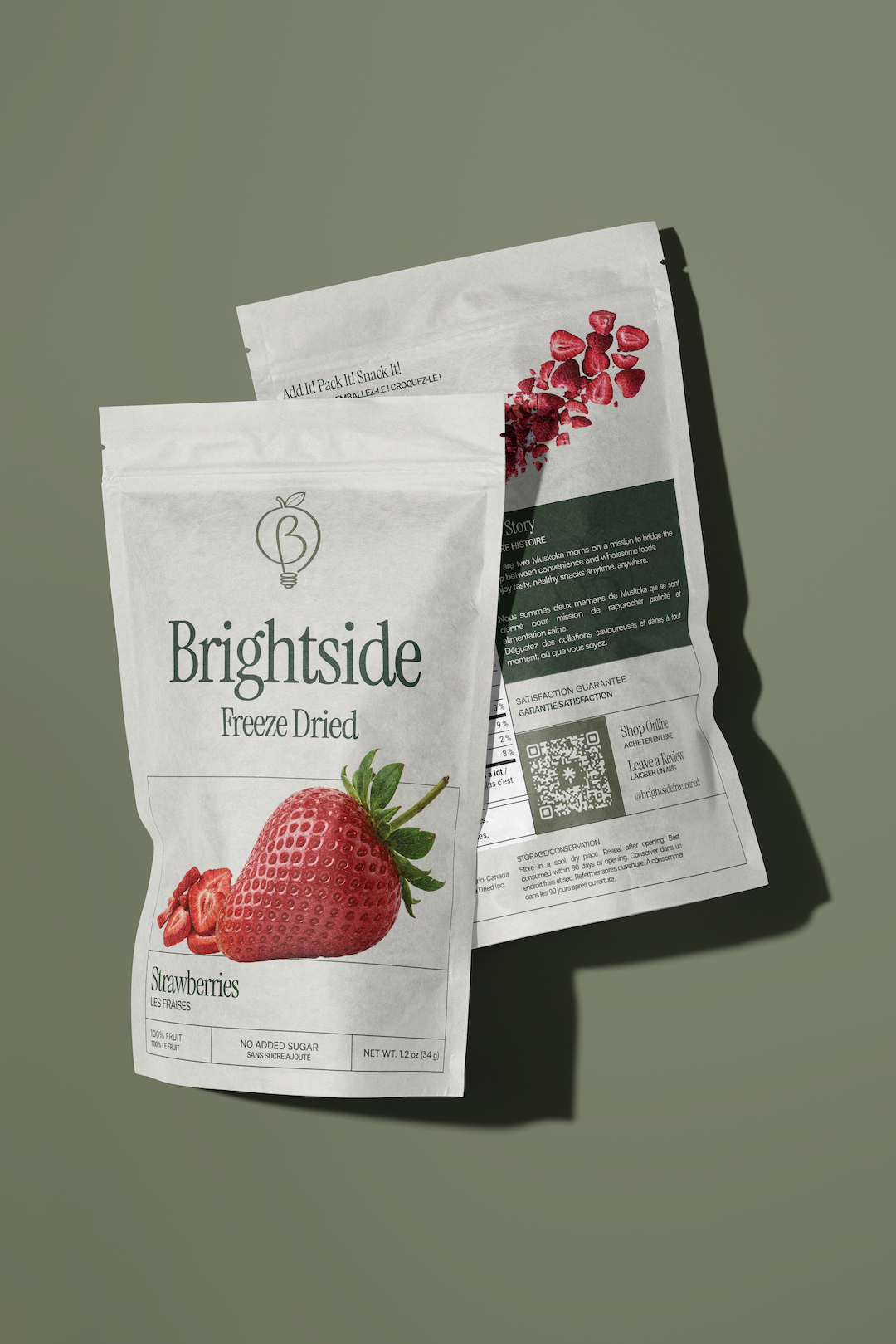Two packages of Brightside Freeze Dried Strawberries on a green surface, one front-facing with a strawberry illustration and the other showing the back of the packaging with product information and a QR code.