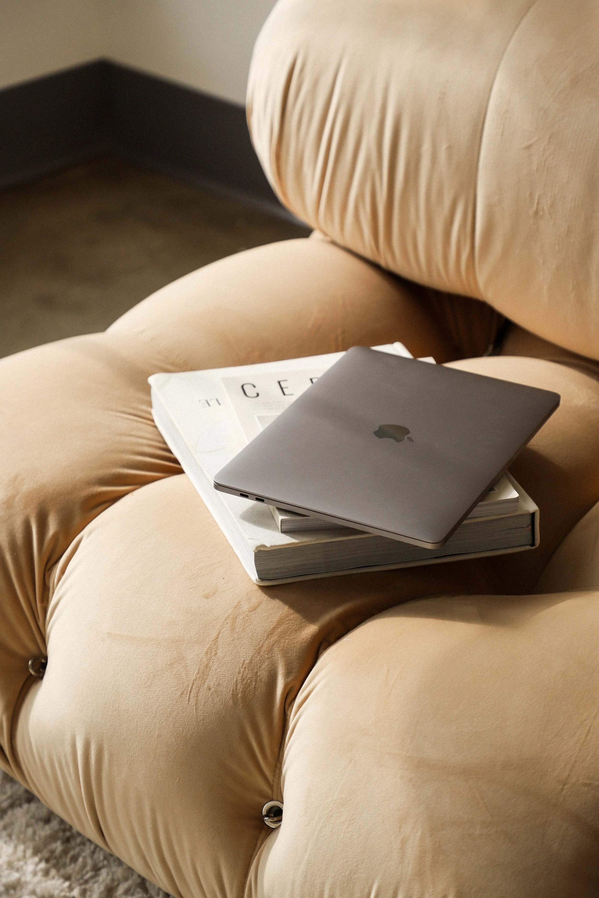 A beige tufted ottoman with a closed silver MacBook and a white magazine resting on top.