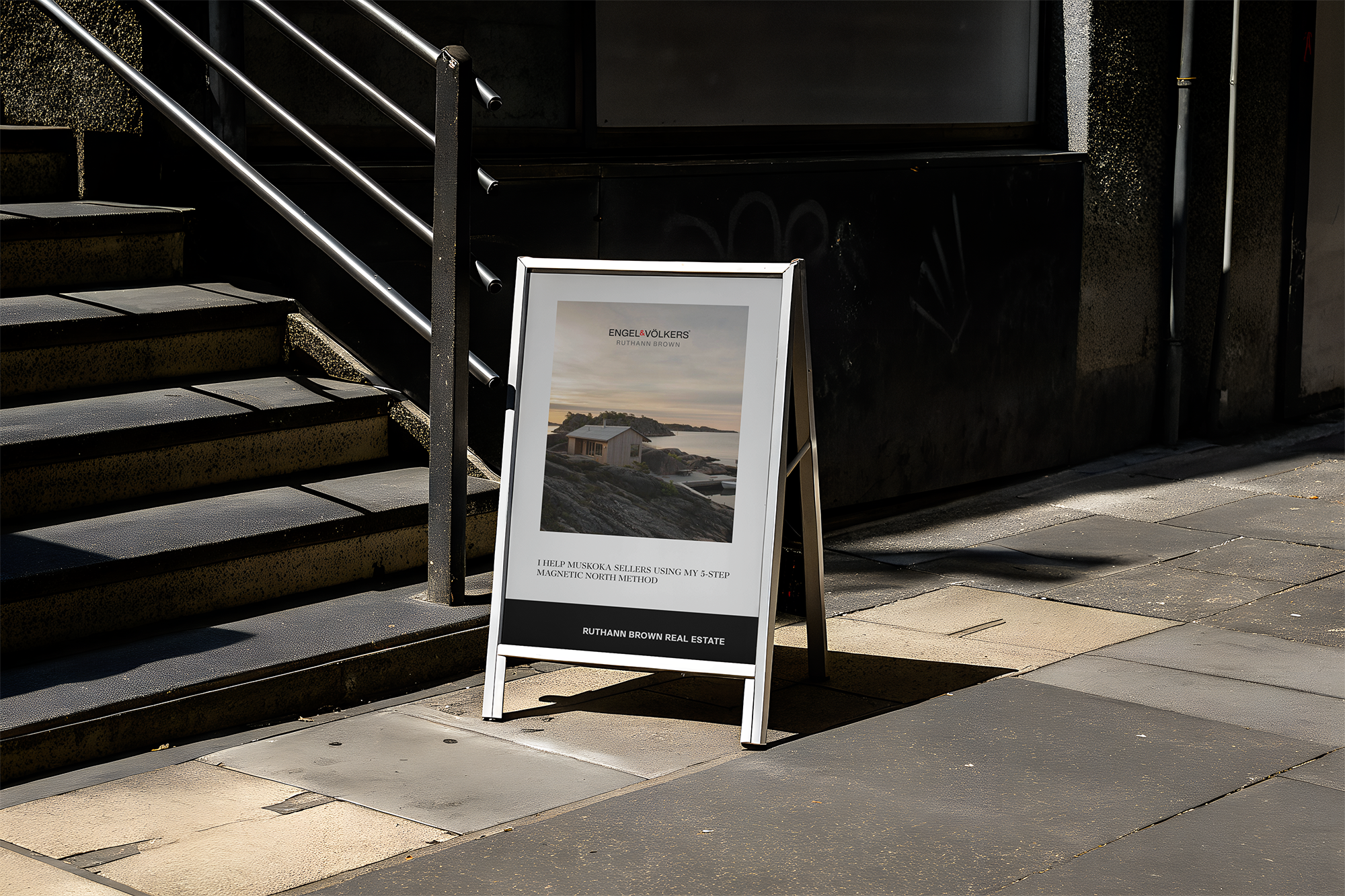 A black staircase with metal railings, leading up to a building entrance. An A-frame sign displays an advertisement for Ruthann Brown Real Estate with a photo of a house and some text, next to the stairs.