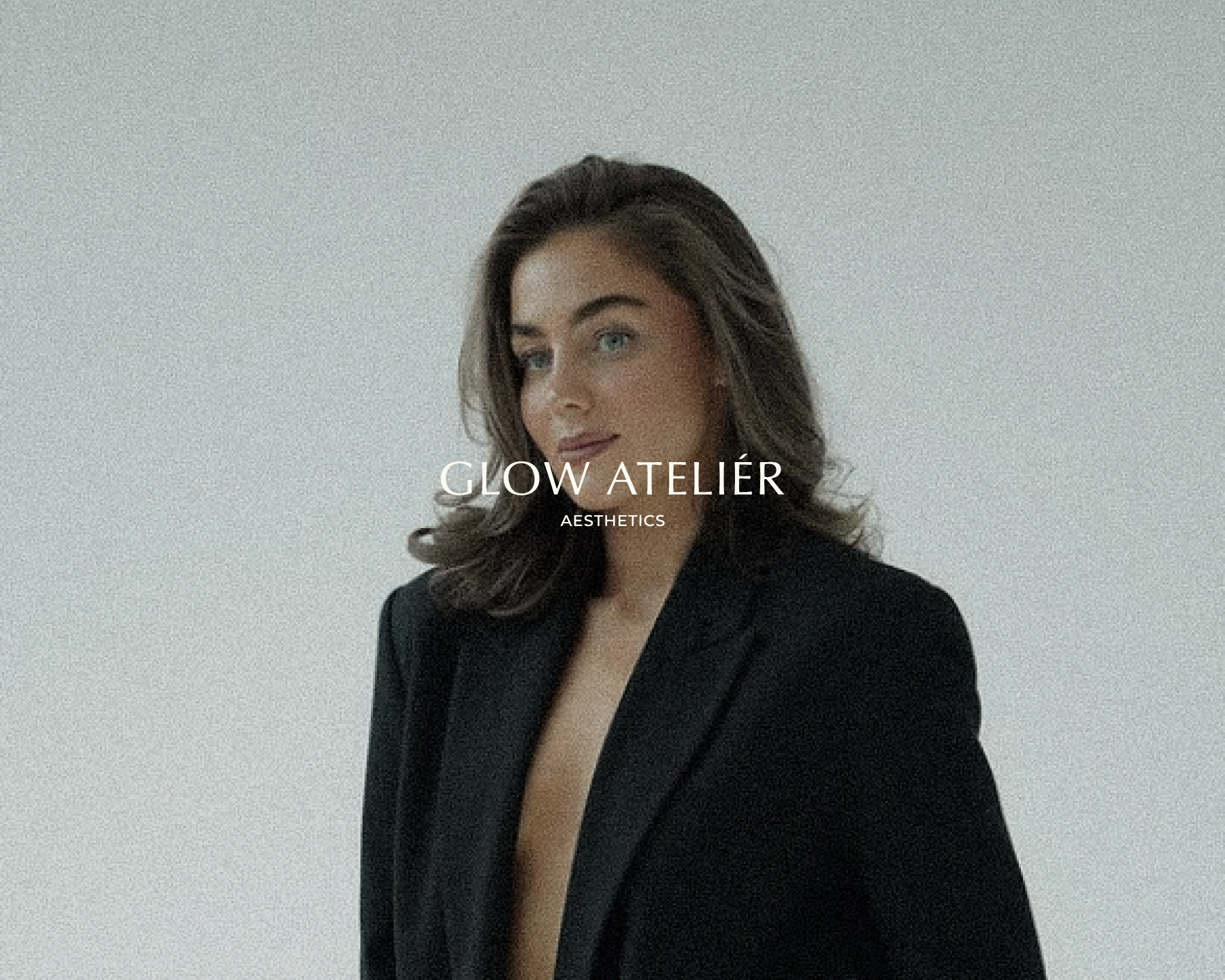 A woman with long wavy brown hair and blue eyes wearing a black blazer with a deep neckline, standing against a plain light gray background.