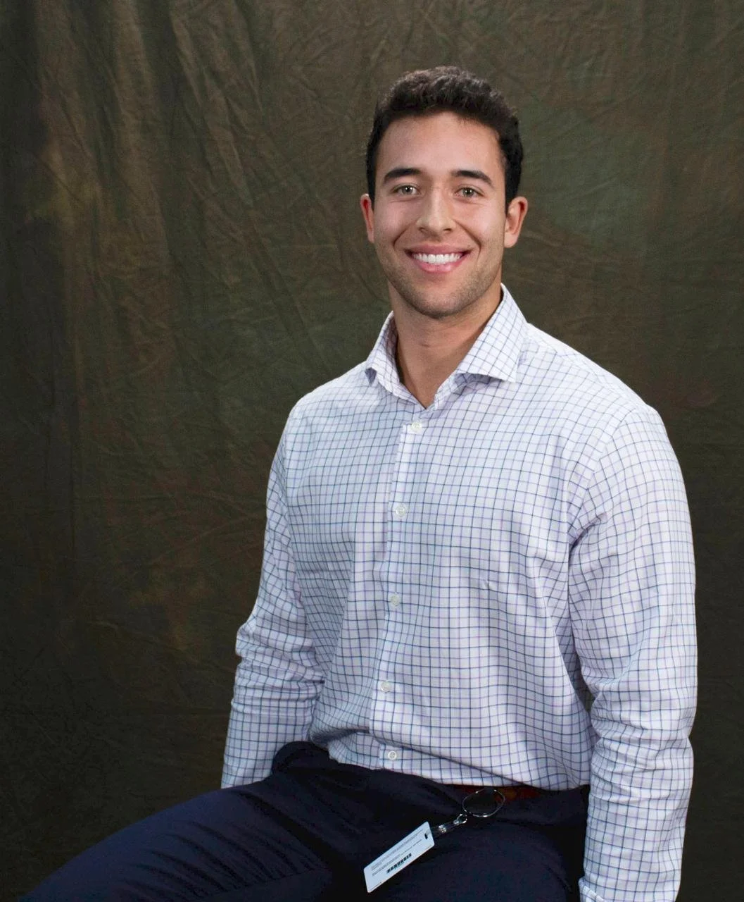 A smiling young man in business attire sitting against a dark backdrop.