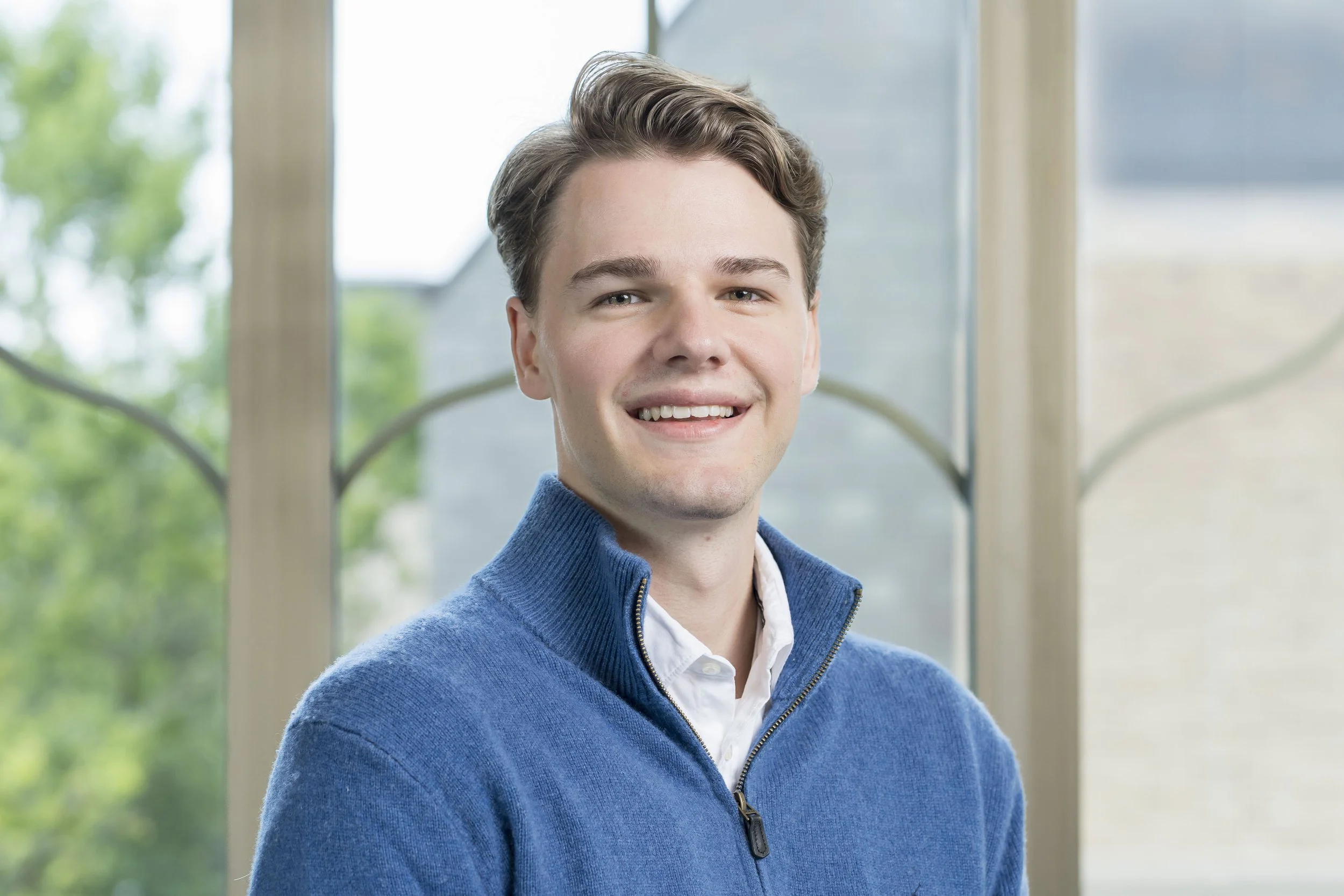 A young man with light skin, brown hair, and blue eyes smiling at the camera. He is wearing a blue zip-up sweater over a collared shirt, standing indoors near a window with greenery outside.