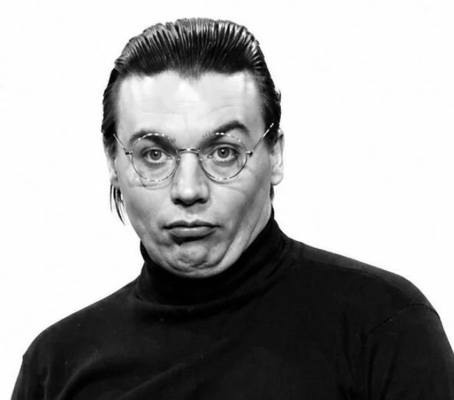 Black and white photo of a man with slicked back hair, round glasses, wearing a black turtleneck, making a humorous facial expression with raised eyebrows and pursed lips.