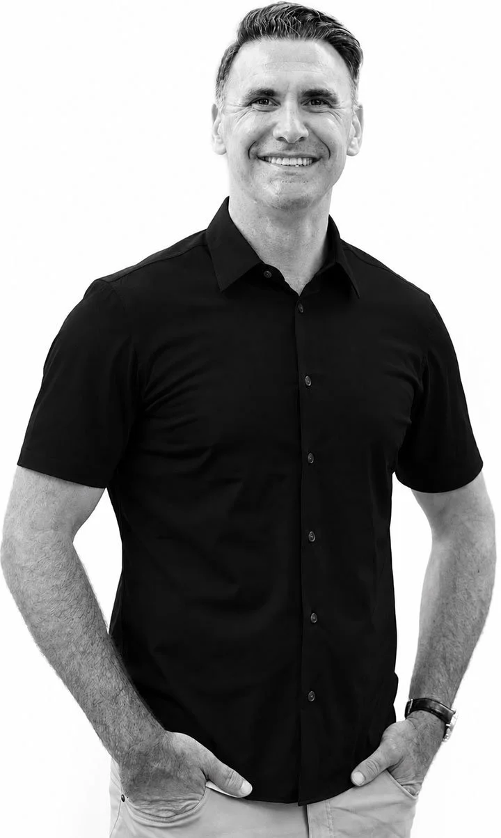 A man smiling, wearing a black button-up shirt and beige pants, with his hands in his pockets, standing against a white background.