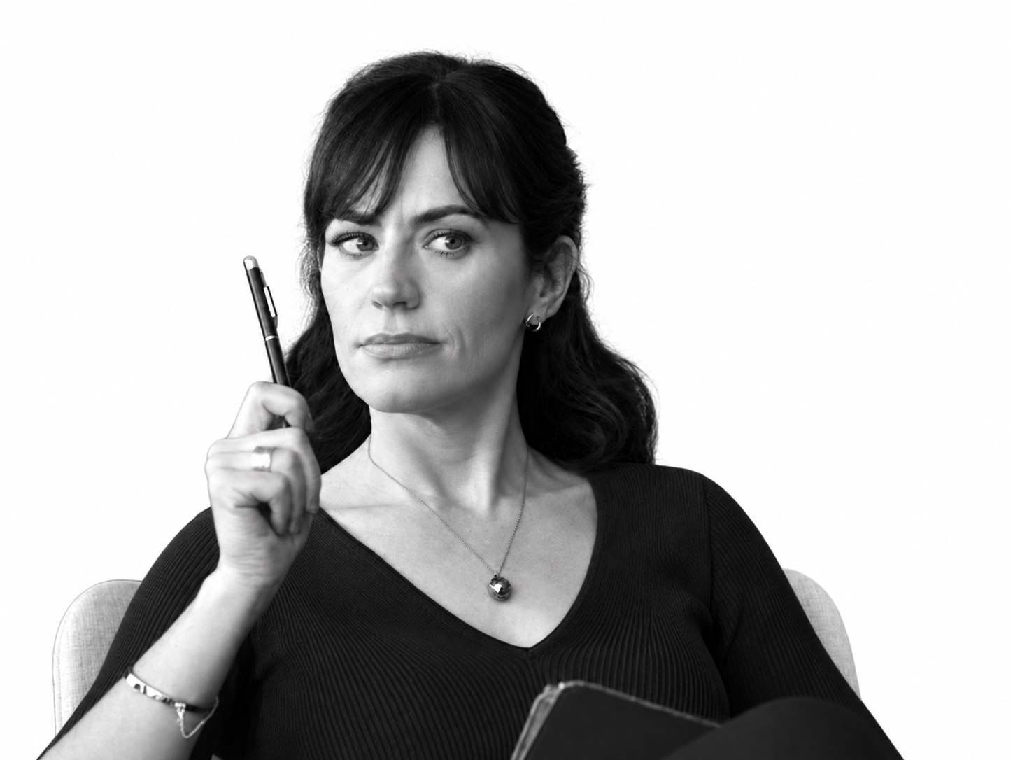 Black-and-white photo of a woman with dark hair holding a pen near her temple, sitting in a chair, wearing a black top and a necklace with a pendant, looking to her left