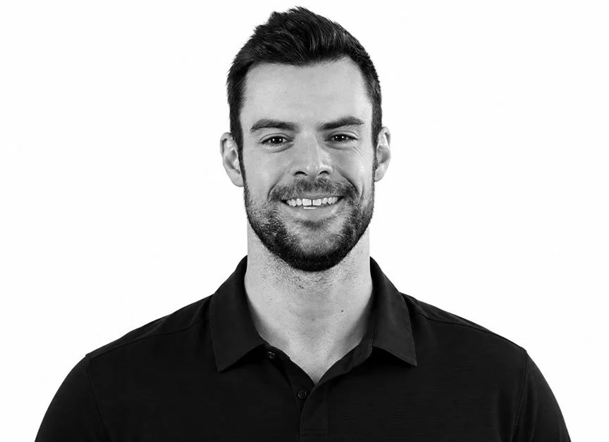 Black and white headshot of a smiling young man with short dark hair and a beard, wearing a black collared shirt against a plain white background.