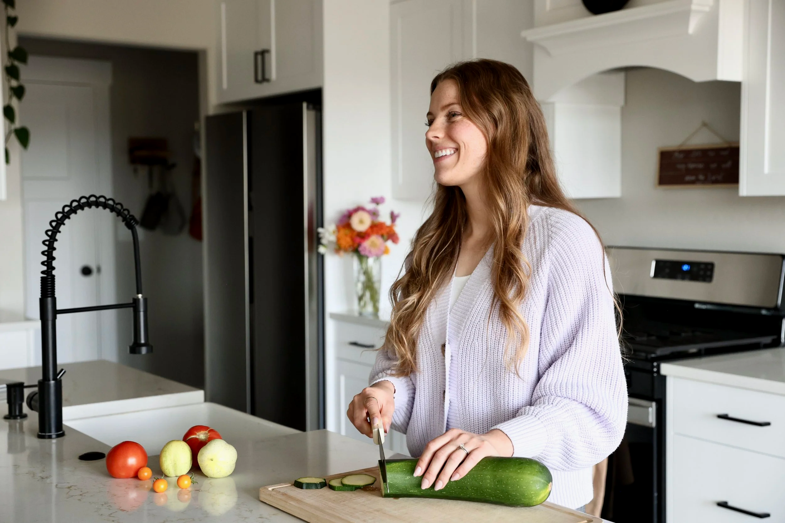 A woman with long red hair smiles while slicing a cucumber on a cutting board in a modern kitchen. There are tomatoes, apples, and cherry tomatoes on the white countertop. A vase with pink, white, and orange flowers is in the background.