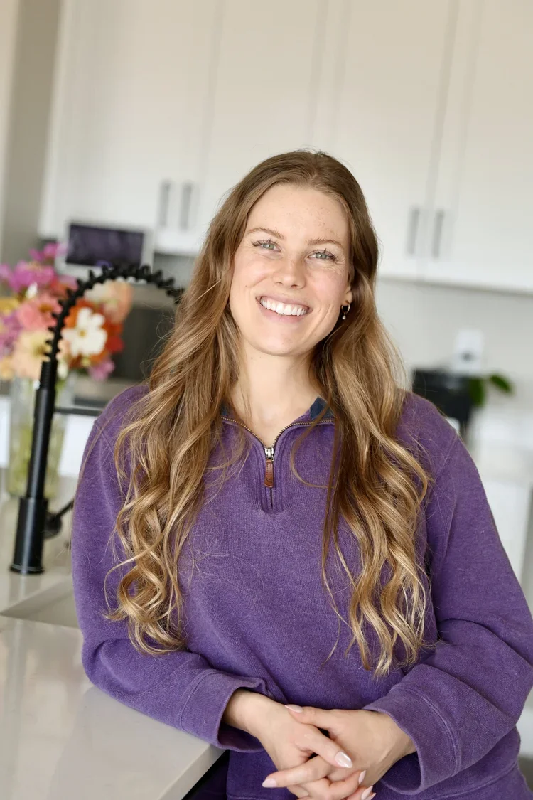 A young woman with long wavy hair, smiling, standing in a kitchen with white cabinets, flowers in a vase, and a black faucet in the background.