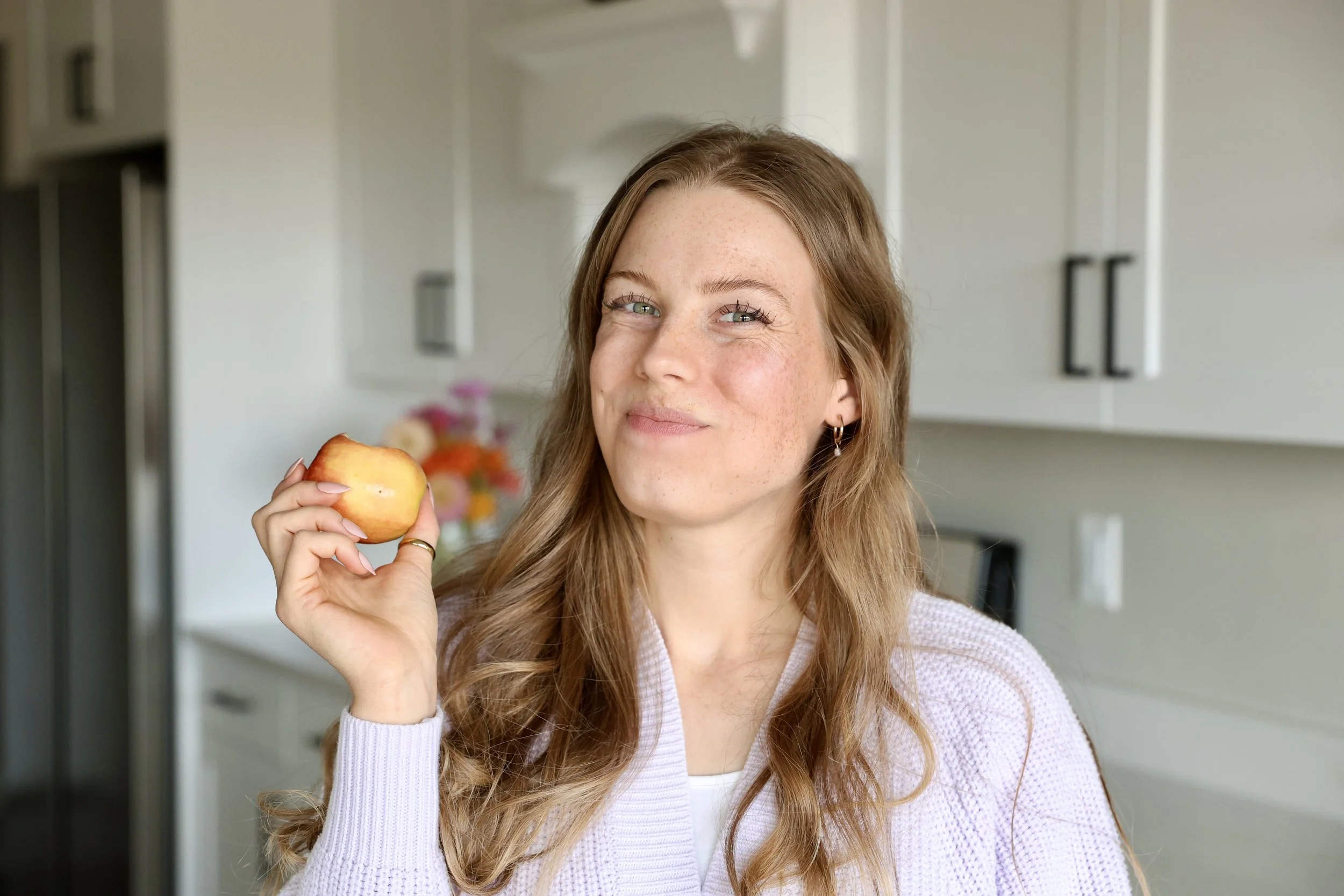 A woman with long, wavy red hair and light skin, holding a red apple near her face, smiling softly in a bright kitchen with white cabinets and a blurred bouquet of flowers in the background.