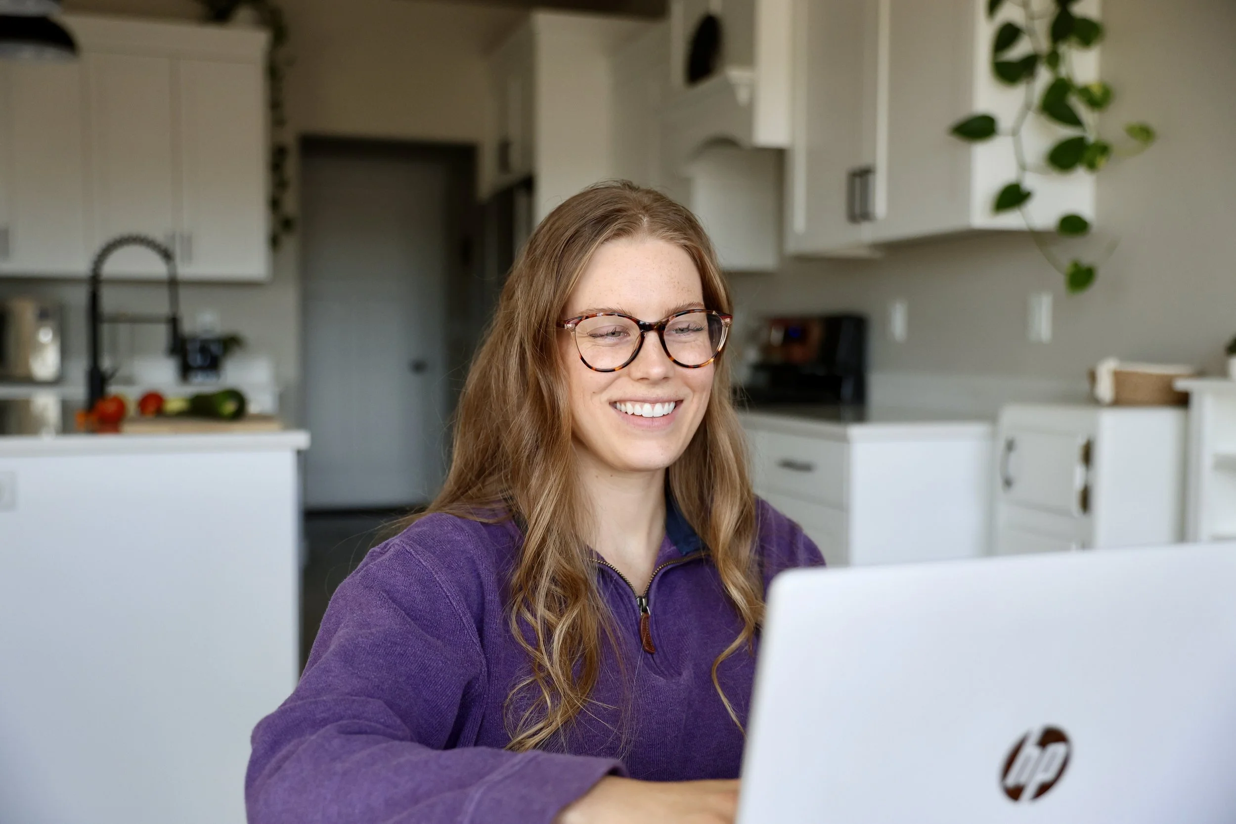 A young woman with long red hair, wearing glasses and a purple jacket, smiling while looking at her laptop screen in a bright kitchen.