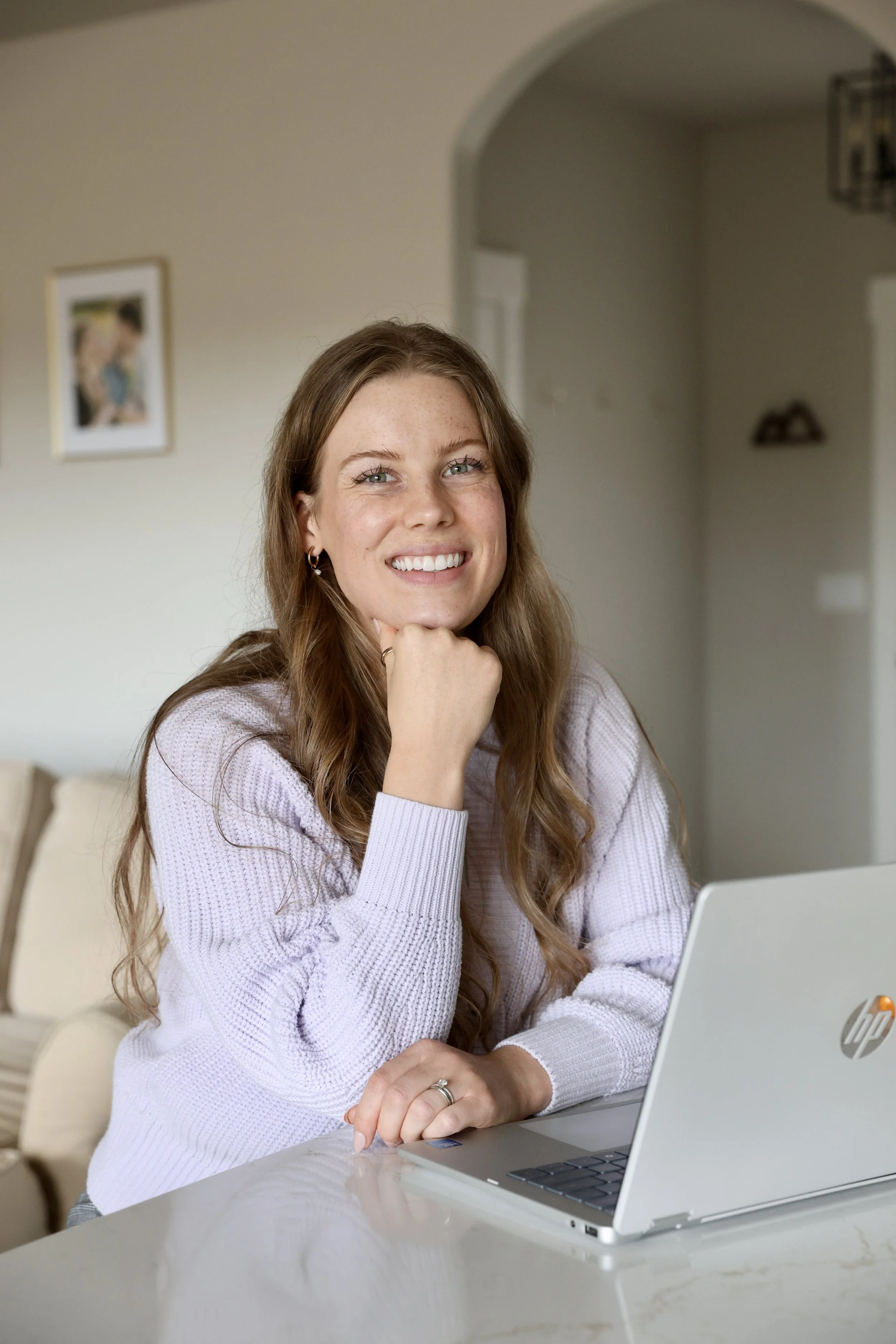 A young woman with long red hair, wearing a light purple sweater, smiling and resting her chin on her hand, sitting at a table with an open HP laptop in front of her in a home setting.