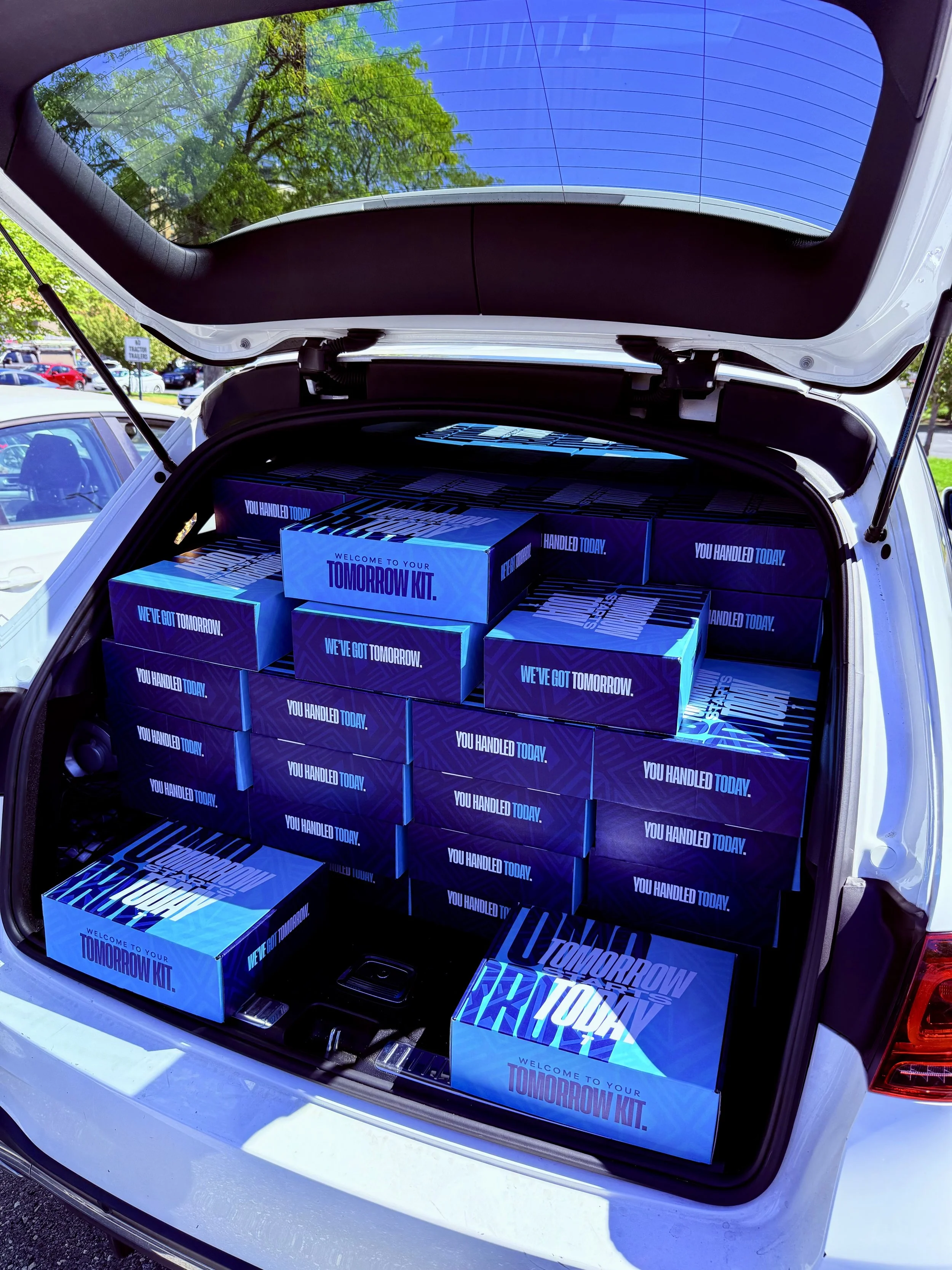 The trunk of a white car filled with multiple blue boxes labeled 'Welcome to your tomorrow kit. We've got tomorrow. You handled today.' and the open hatch showing the car's taillights and a partly cloudy sky with some green trees reflection.