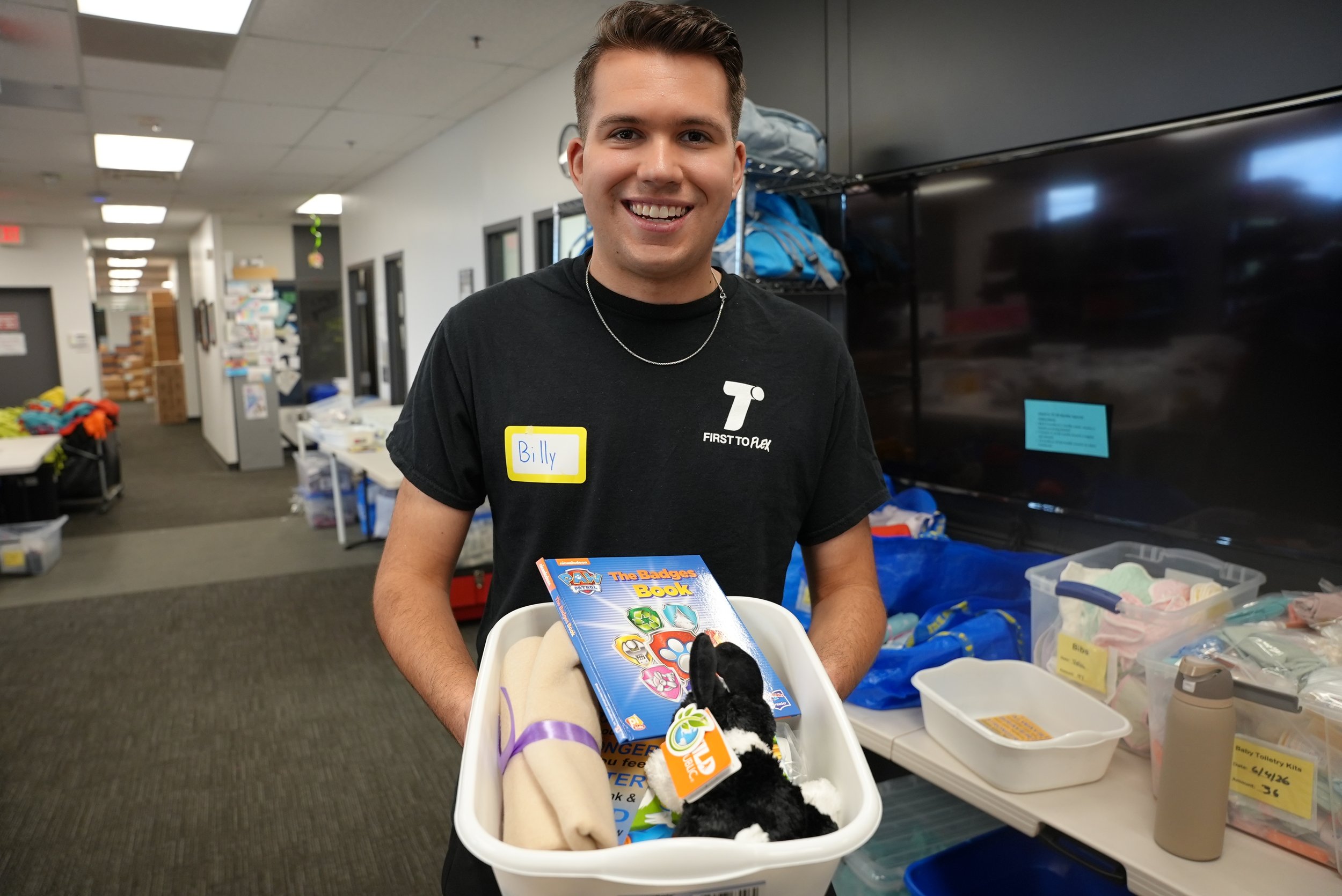 A young man with short brown hair, smiling and wearing a black t-shirt and a silver chain, stands in an indoor setting holding a large container with stuffed animals and a book titled "The Badges Book". He has a yellow name tag that says Billy. The background shows shelves with blue backpacks, cubbies, and various supplies.