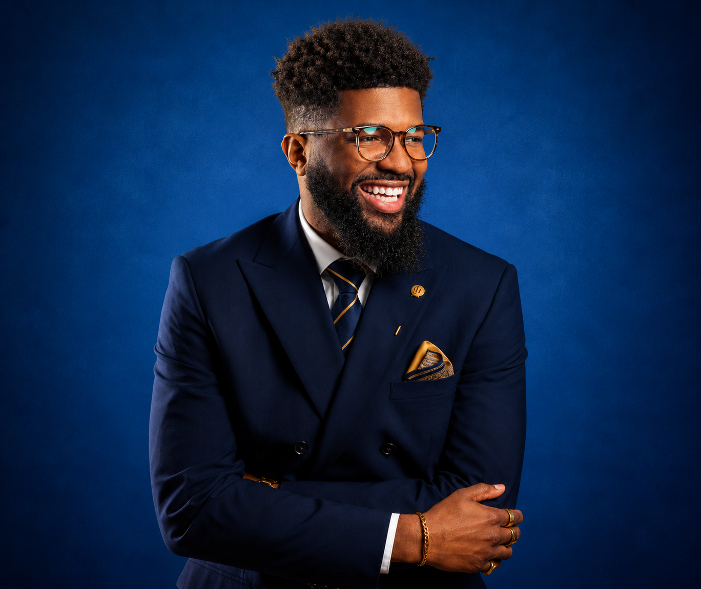 A smiling man with glasses and a beard dressed in a navy blue suit with a tie and a pocket square, standing against a blue background.