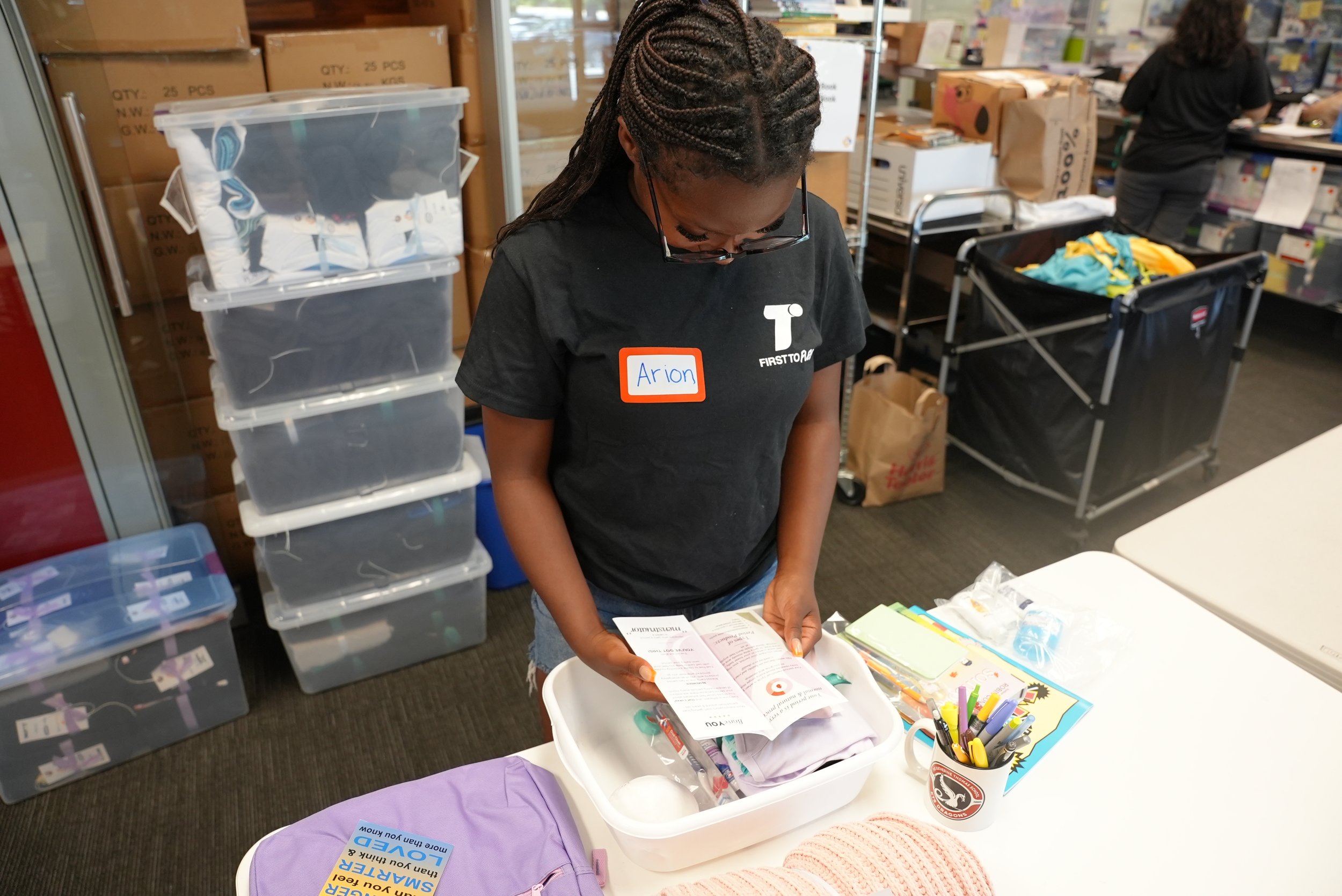 A woman with braided hair and glasses, wearing a black shirt with a name tag that says 'Arion,' is reading a pamphlet or brochure at a cluttered table. The background shows shelves with plastic storage bins, cardboard boxes, and other people working or organizing in a room.