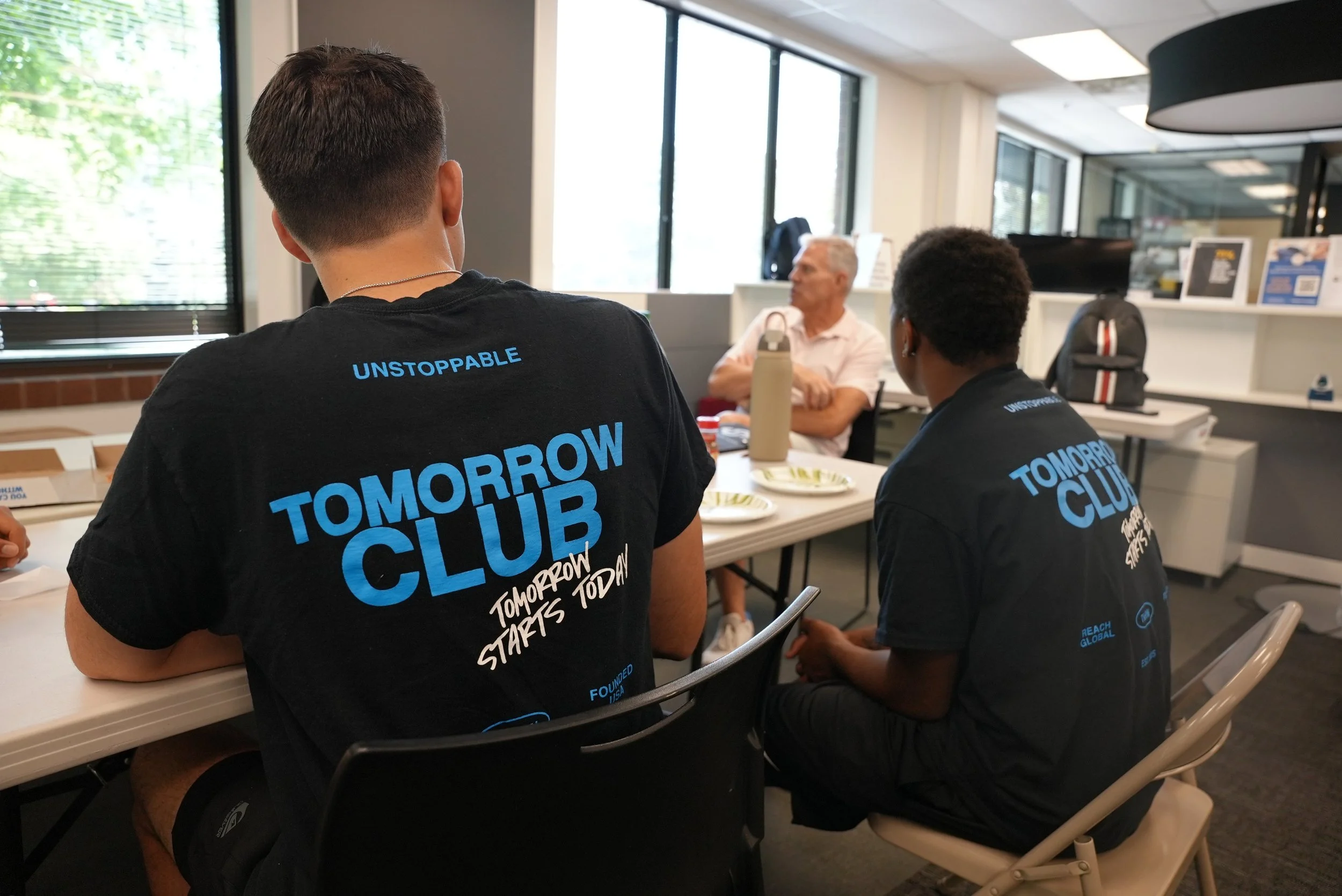 Two young individuals sitting at a table wearing black T-shirts with blue text that reads 'Tomorrow's Club' and additional text. They are in a room with large windows, papers, and items on the table. An older man with gray hair is seated across from them. The room has shelves with pictures and materials, and the setting appears to be a community meeting or group activity.