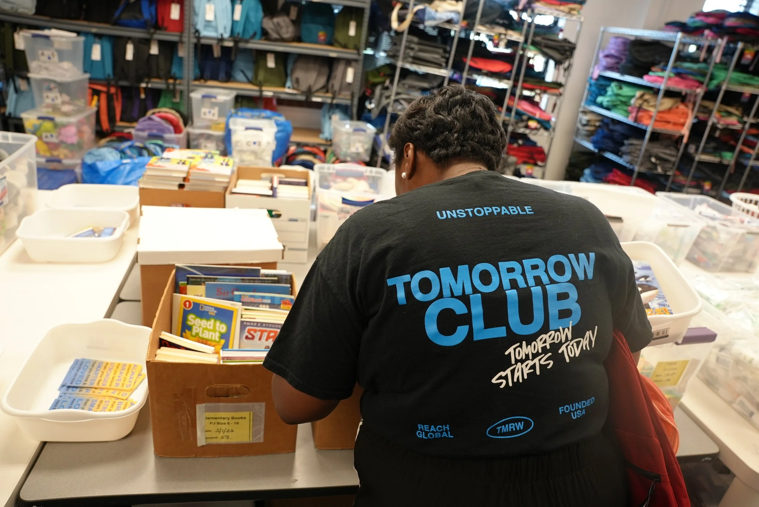 Person with short curly hair wearing a black T-shirt with blue text, sitting at a table in a room with shelves filled with colorful backpacks and bins. The T-shirt reads 'UNSTOPPABLE' and 'TOMORROW STARTS TODAY'.