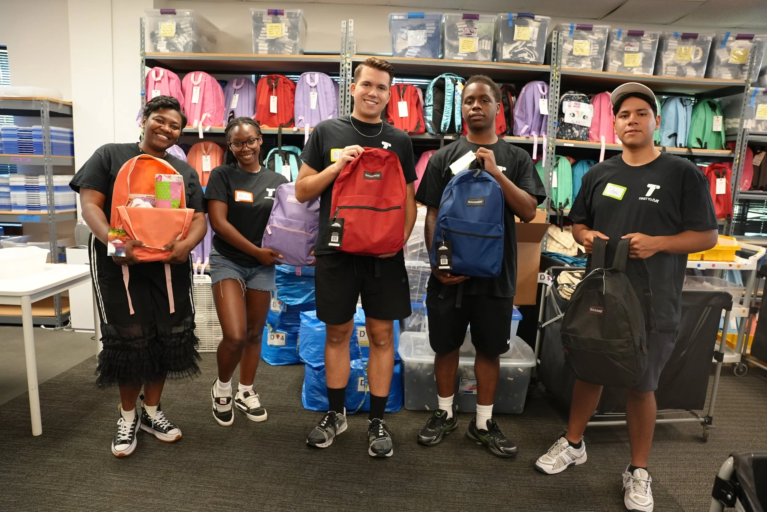 A group of five young people holding backpacks and standing in front of shelves filled with colorful backpacks and storage bins in a room, possibly a warehouse or donation center.