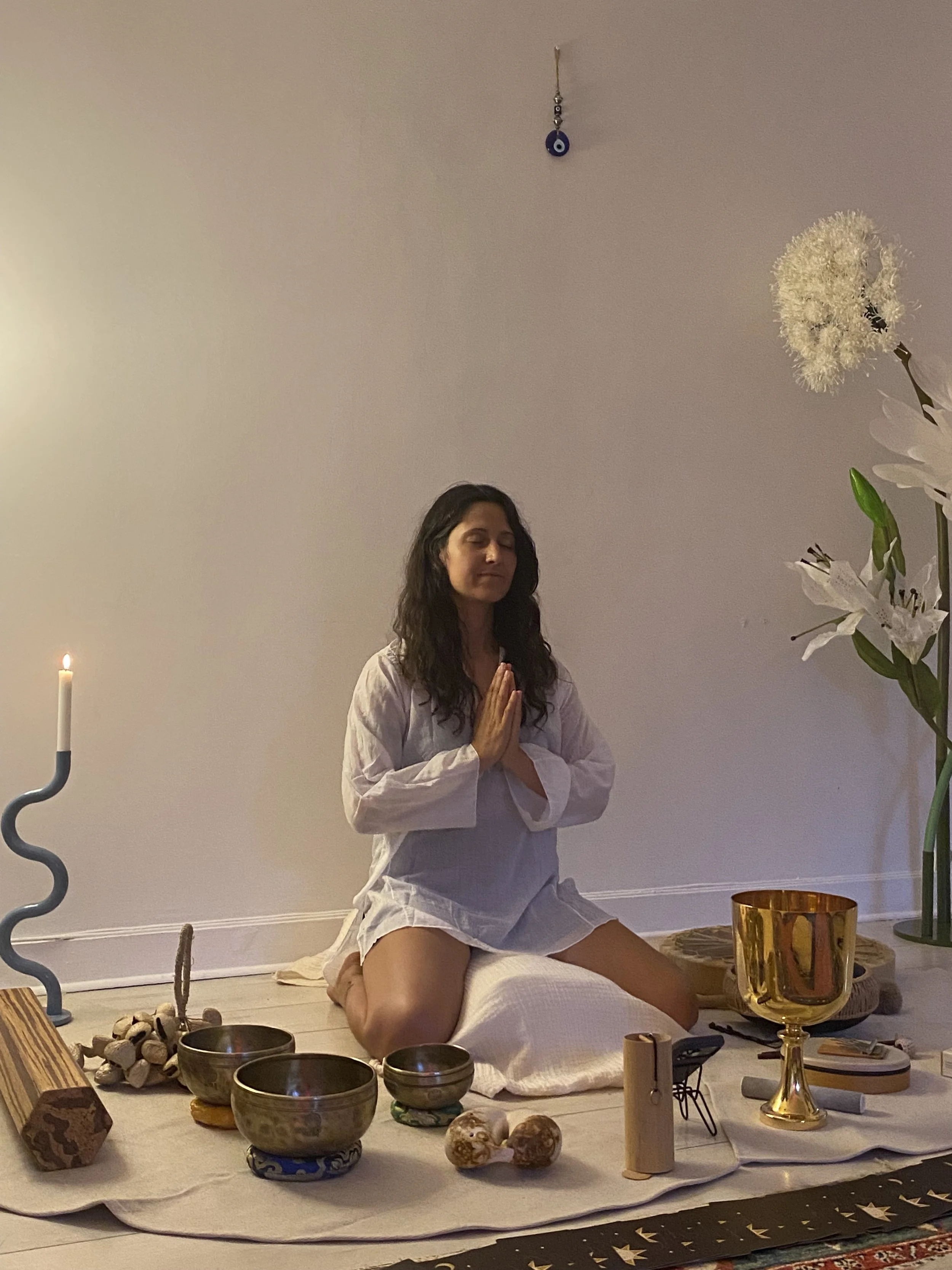 Une femme faisant de la méditation ou de la prière en position de lotus, assise sur un tapis, entourée d'objets décoratifs et rituels sur le sol, dans une ambiance calme avec une lumière douce.
