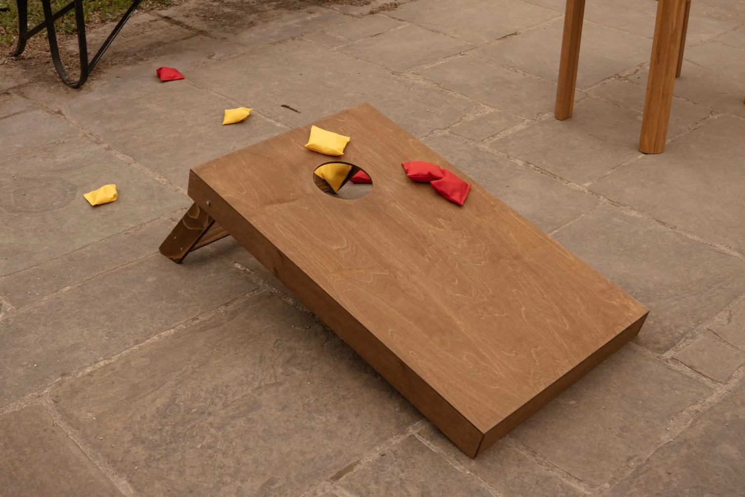A wooden cornhole board with some red and yellow bean bags on and around it, on a stone patio.