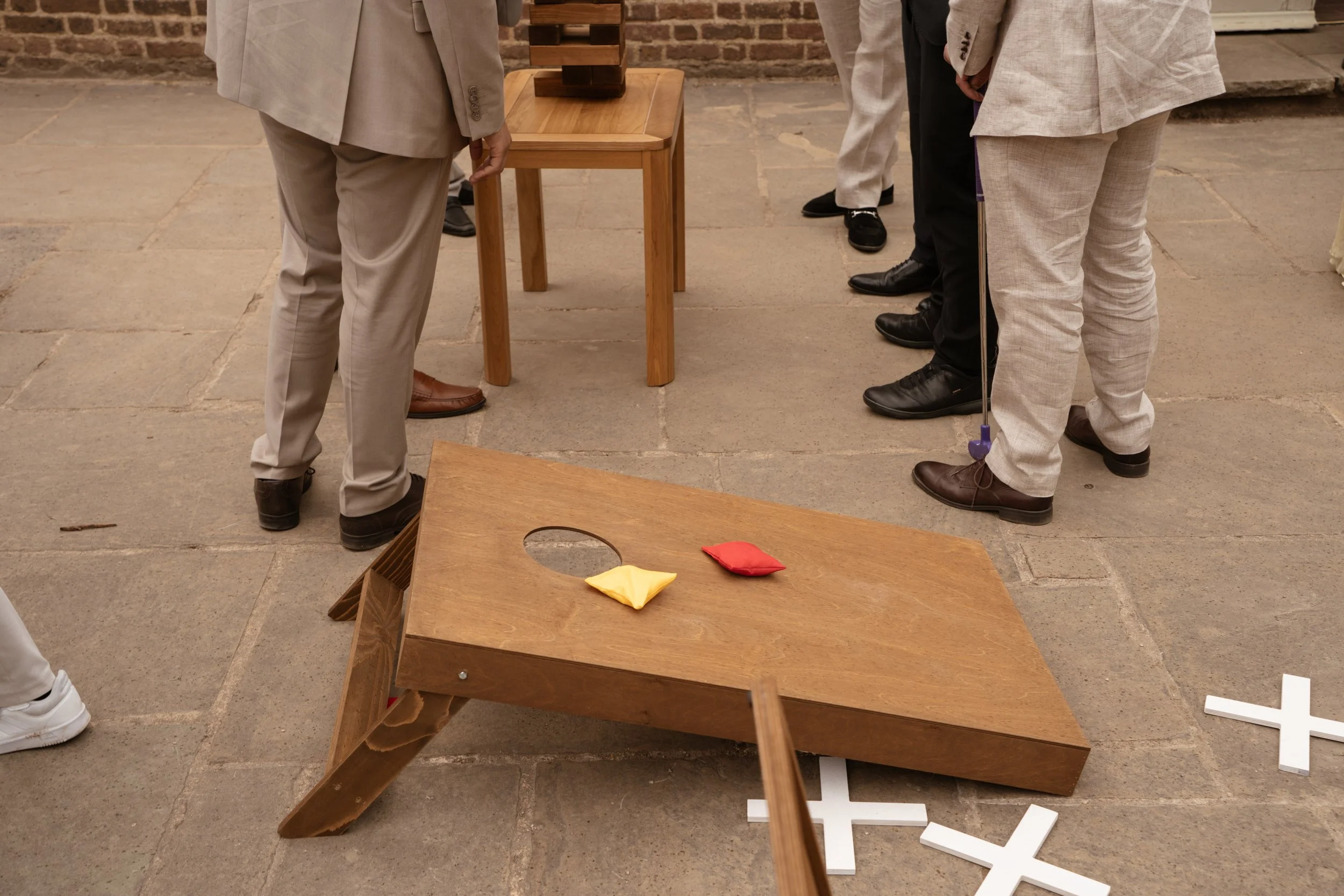 People playing a game of Jenga outdoors, with a wooden Jenga tower on a small table and colored fabric bags on a wooden game board nearby.
