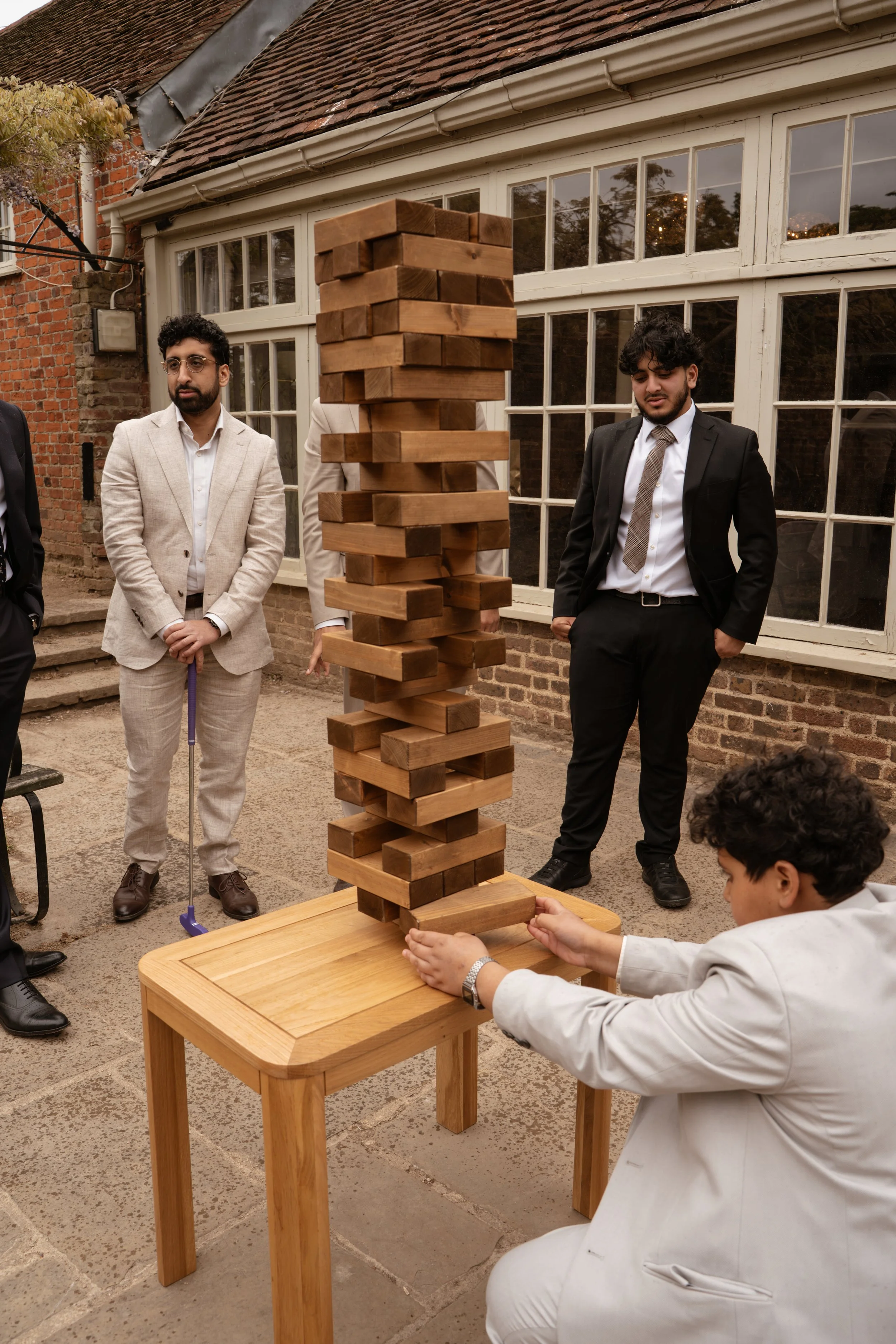A group of people dressed in formal attire playing Jenga outdoors near a brick building with large windows.