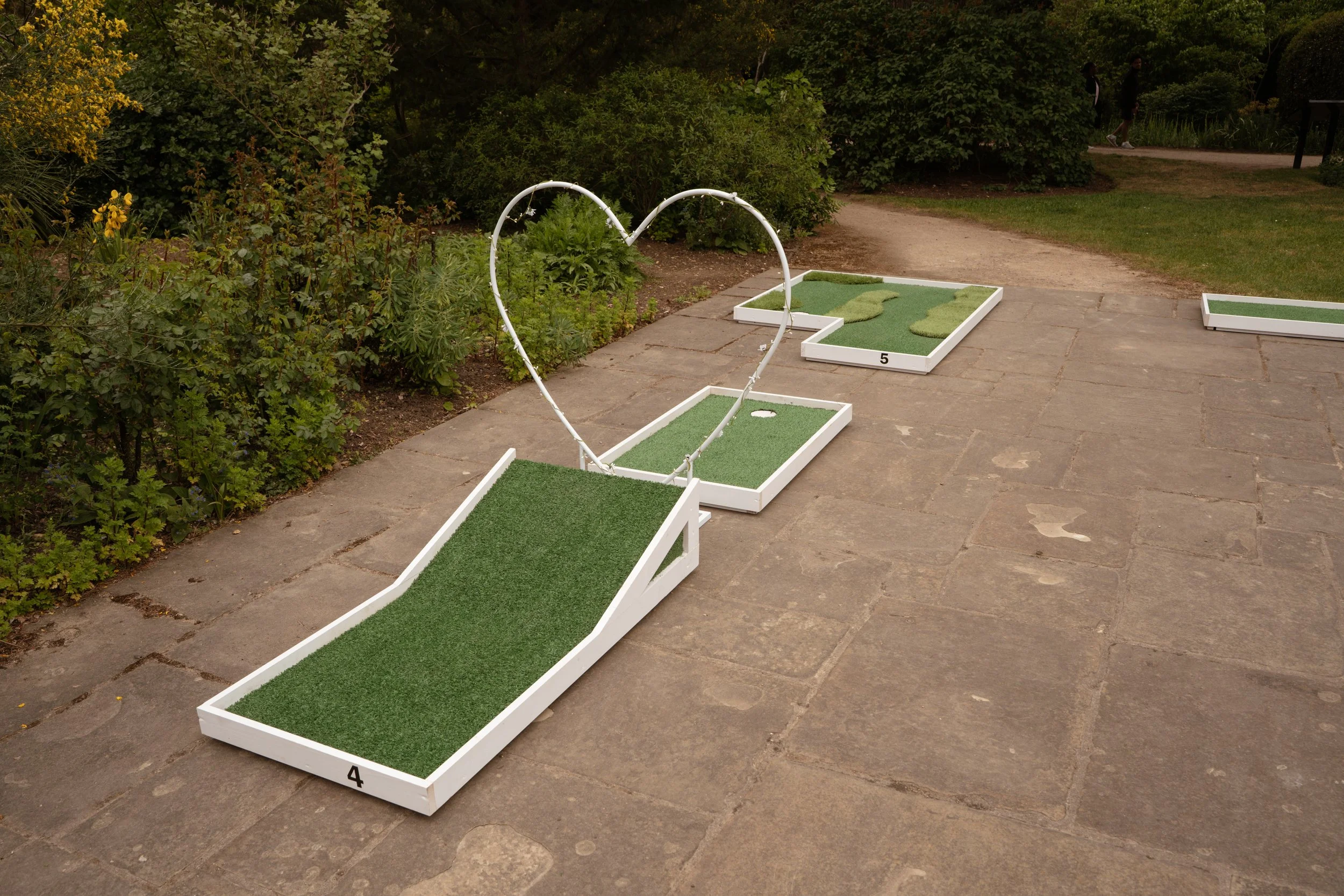 Miniature golf course with holes numbered 4 and 5, featuring artificial turf and white borders, situated on a paved pathway in a garden park with green bushes and trees in the background, and two people walking along a dirt trail in the distance.